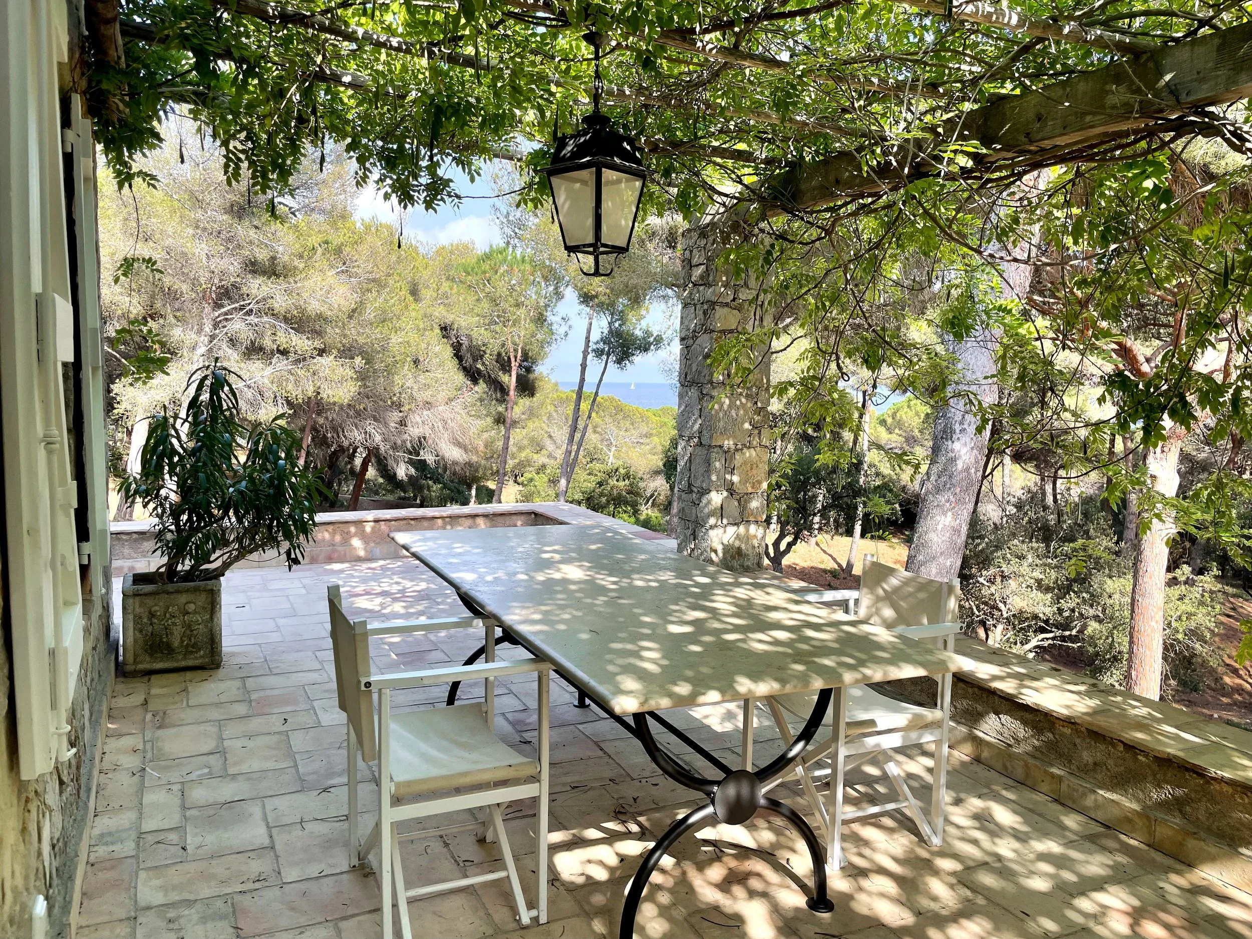 Idyllische mediterrane Terrasse eines Sommerhauses in St. Tropez mit rankenüberwachsener Pergola, Natursteinbelag, Esstisch, Hängelaterne und Blick auf Pinien sowie das Meer.