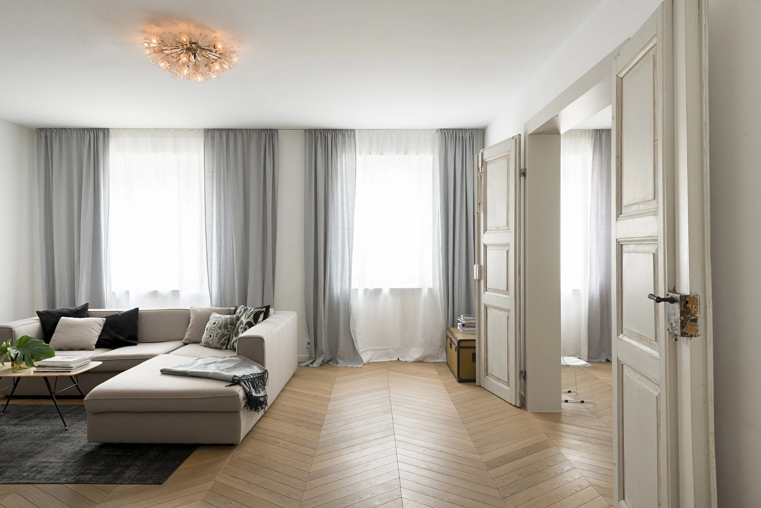 Helles Wohnzimmer mit beigem Ecksofa, grauen Vorhängen, warmem Licht und einer geöffneten, klassischen Altbau-Flügeltür.