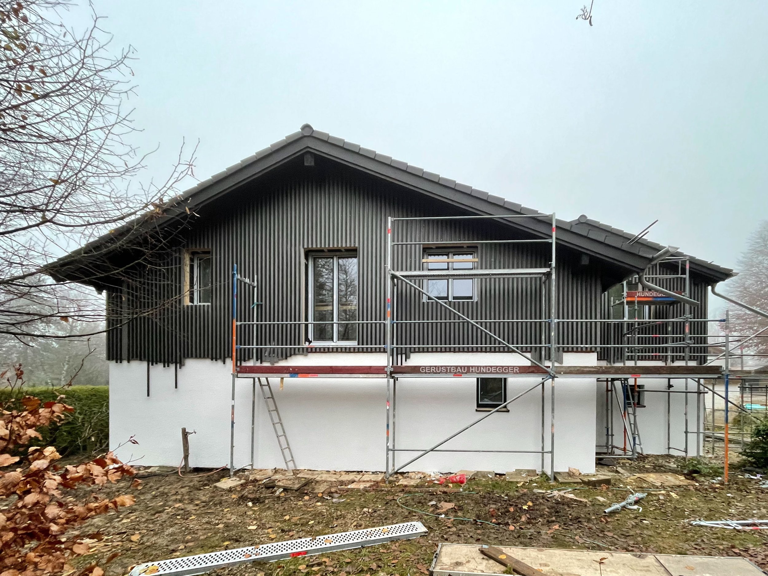 Baustellenfoto eines modernen Einfamilienhauses mit Satteldach, weiß verputztem Erdgeschoss und dunkelgrauer Holzverschalung im Obergeschoss.