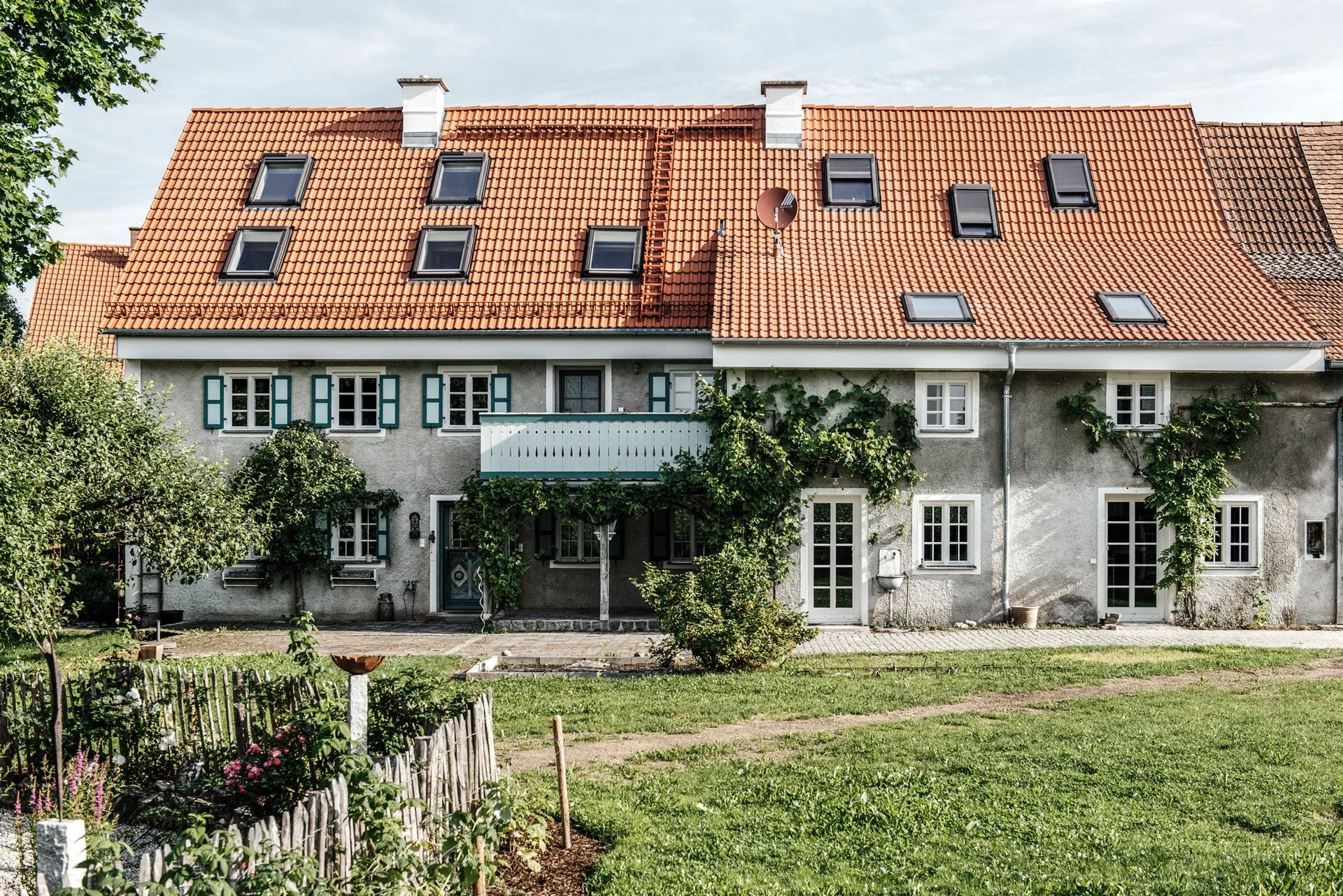 Frontalansicht eines langgestreckten, zweigeschossigen Bauernhauses mit rotem Ziegeleindeckungs-Satteldach, zahlreichen Dachfenstern, grünen Klappläden, Balkon, Fassadenbegrünung und Garten im Vordergrund.