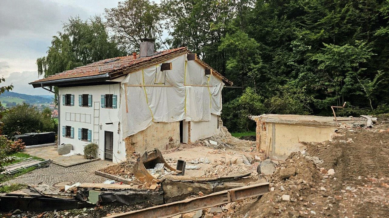 Seitenansicht eines denkmalgeschützten Bauernhauses mit weißer Putzfassade und blauen Fensterläden, dessen Giebel und Innenräume für Umbauarbeiten freigelegt wurden, daneben Erdarbeiten.