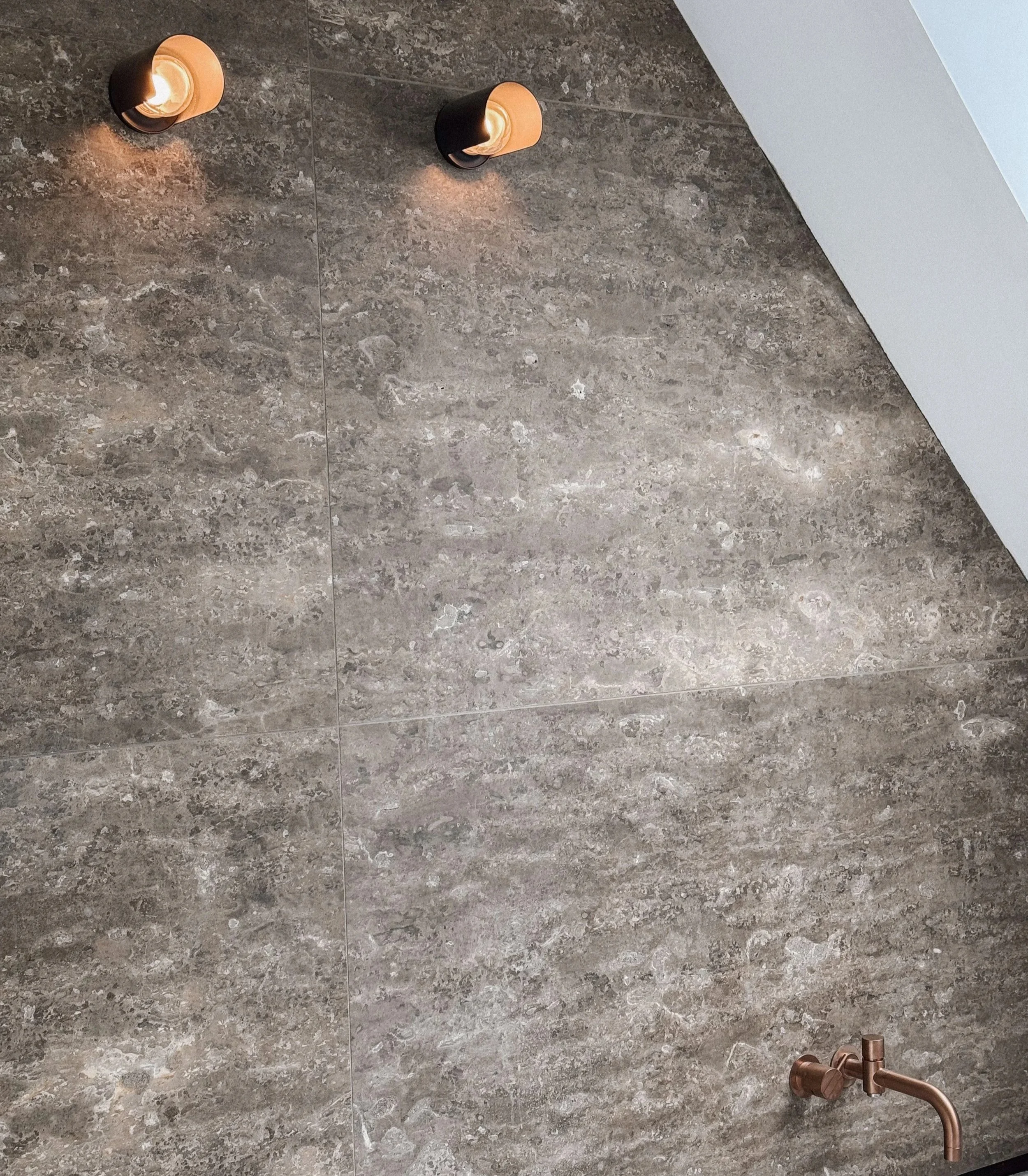 Detailaufnahme einer Badwand aus großformatigen, grau-braunen Natursteinfliesen mit warmen Wandstrahlern und einer kupferfarbenen Unterputz-Armatur.