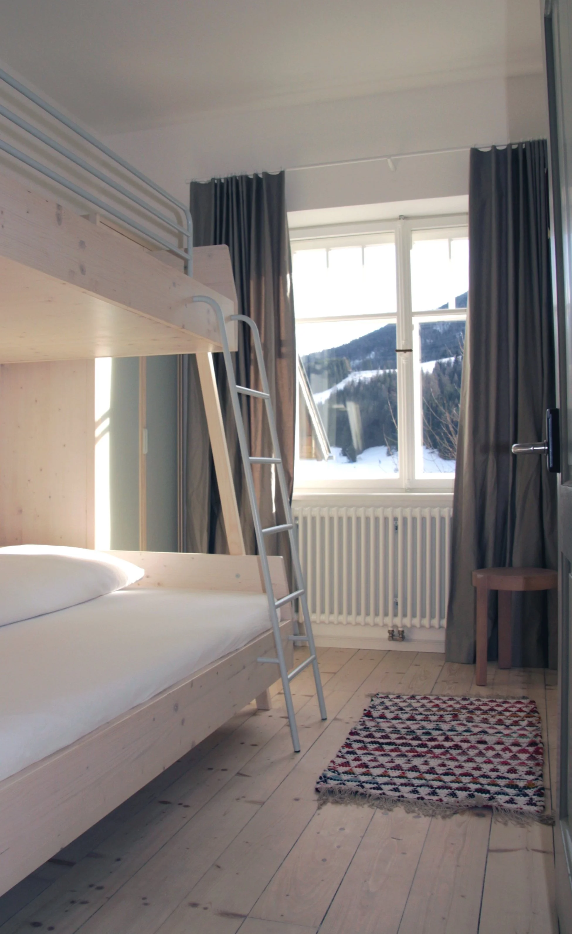 Foto eines hellen Holz-Etagenbetts mit Aluleiter im Hotelzimmer, Dielenboden und Blick durch das Sprossenfenster auf die Berge.