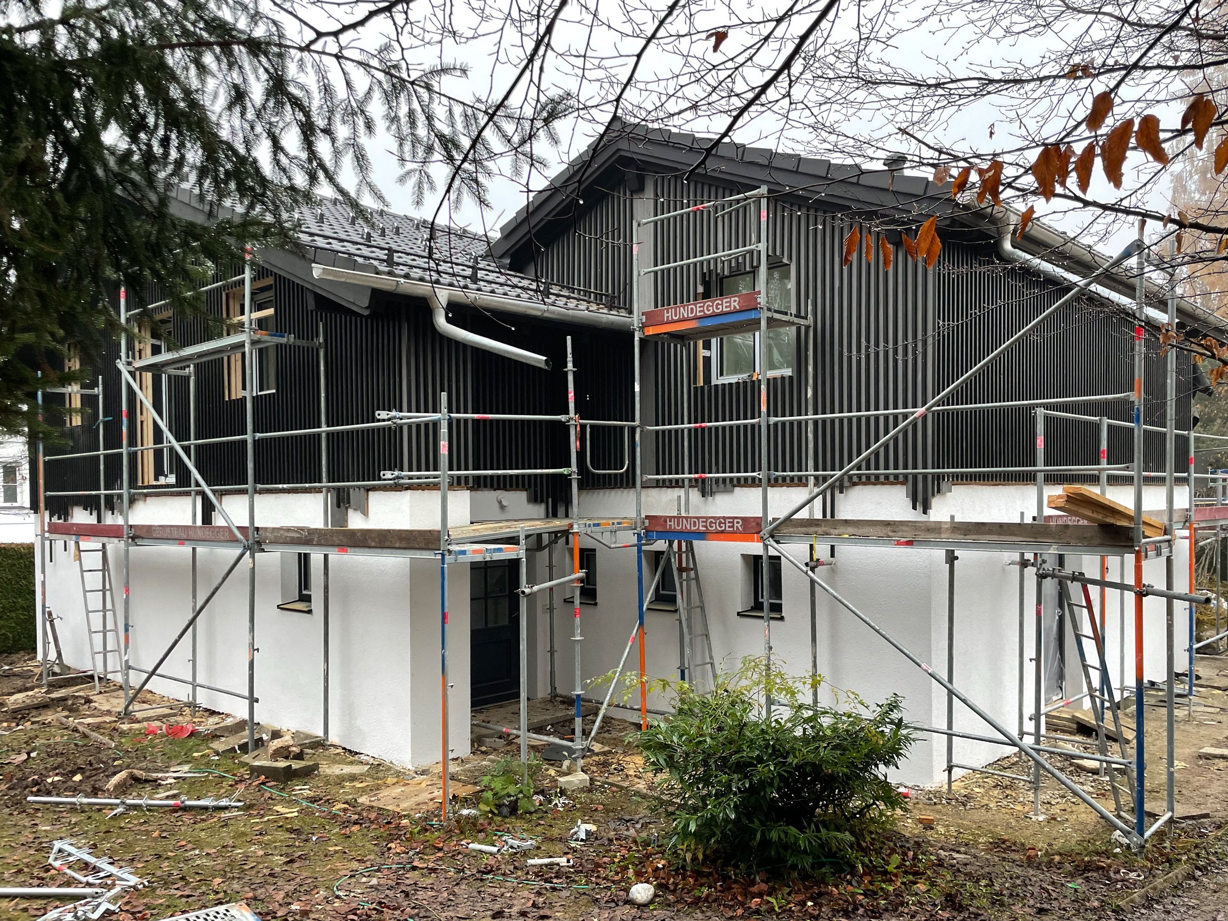 Baustellenfoto eines modernen Einfamilienhauses mit Satteldach, weiß verputztem Erdgeschoss und dunkelgrauer Holzverschalung im Obergeschoss.