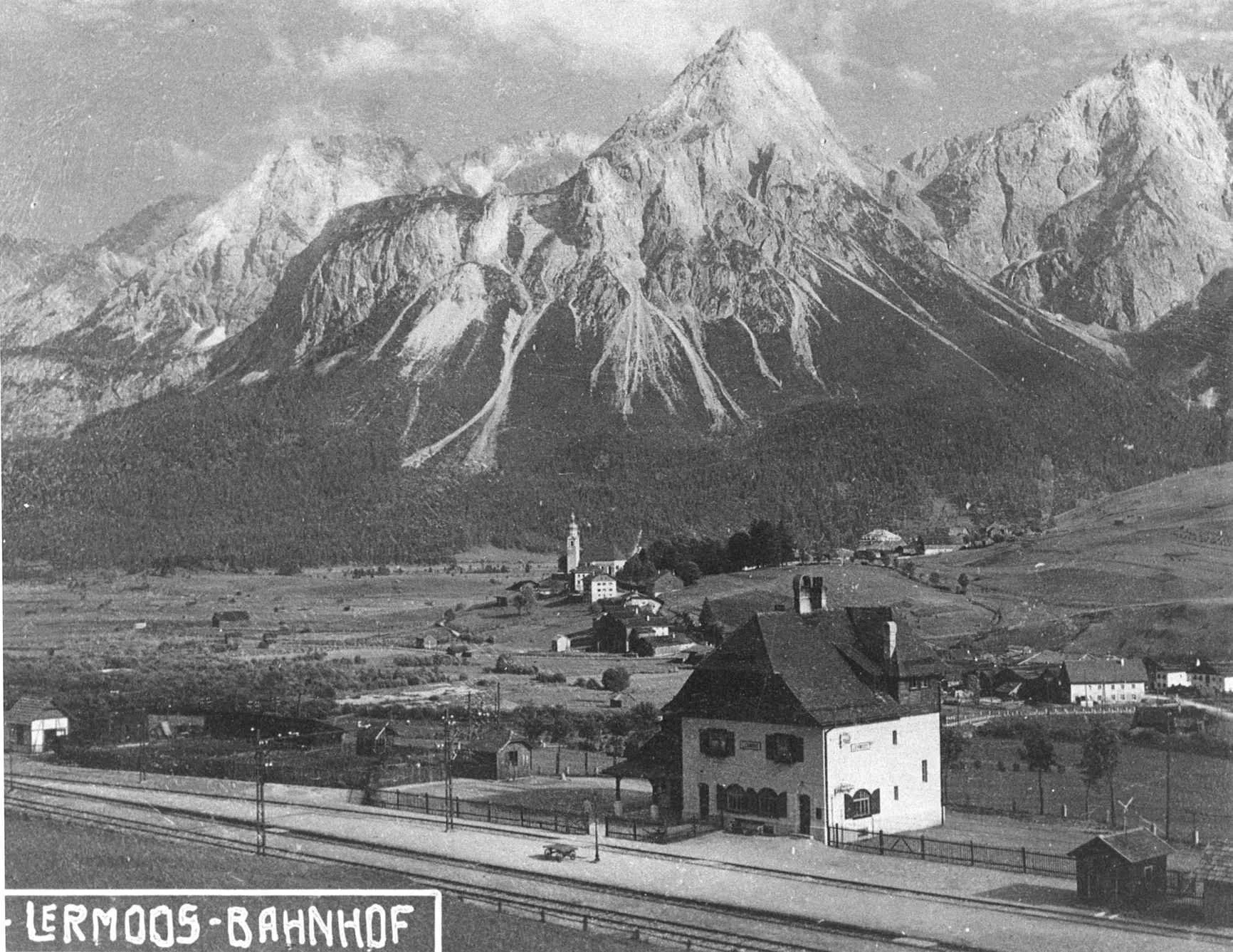 Historisches Foto des Bahnhofs Lermoos im Vordergrund mit Gleisanlagen, dem Dorf Lermoos mit Kirche im Mittelfeld und dem imposanten, felsigen Gebirgsmassiv der Zugspitz Arena im Hintergrund.