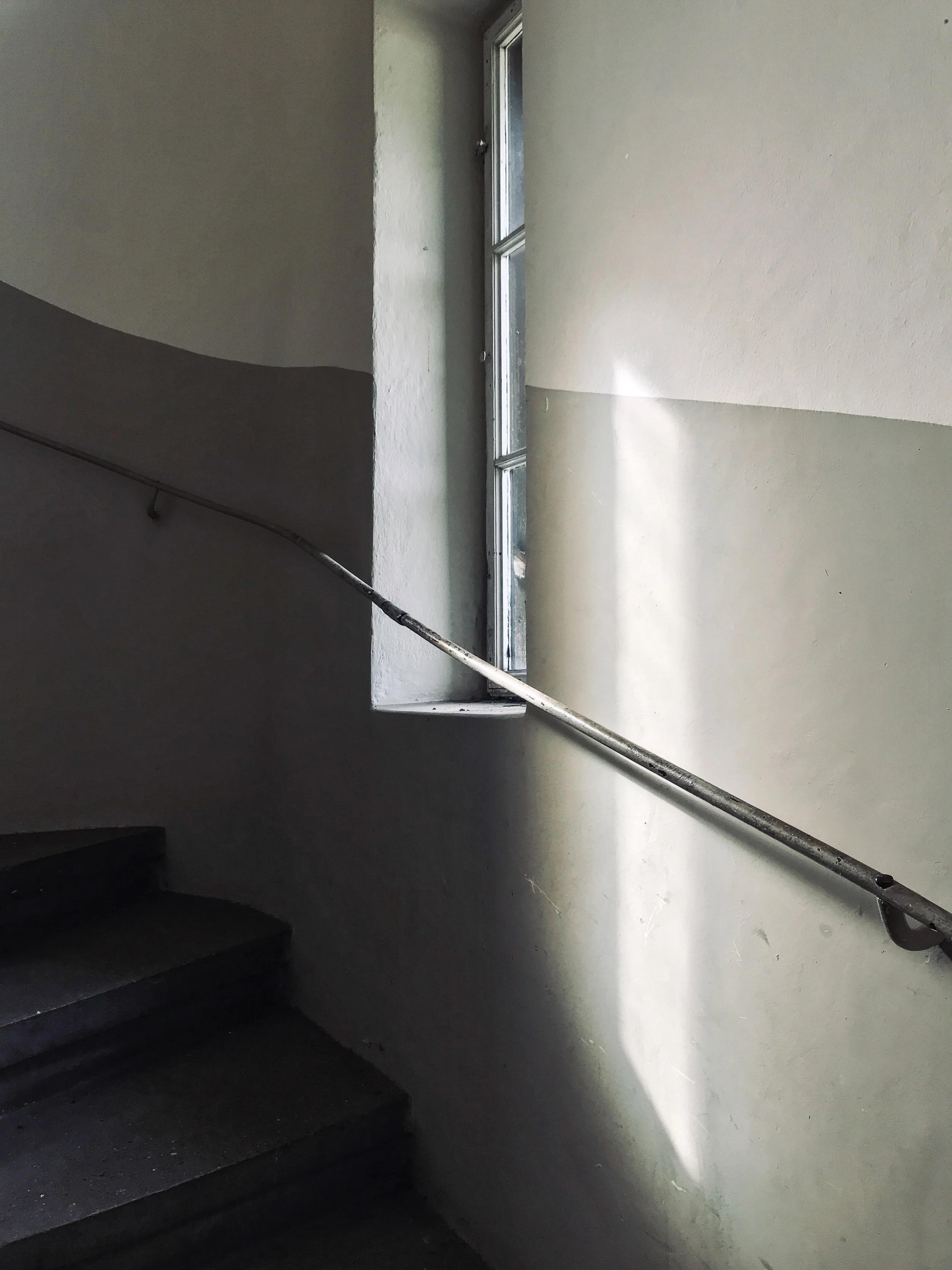 Detailaufnahme im historischen Treppenhaus des Bahnhofs Lermoos mit zweifarbiger Wandgestaltung, filigranem Metallhandlauf, Massivholztreppe und sonnigem Lichteinfall durch ein tiefes Sprossenfenster.