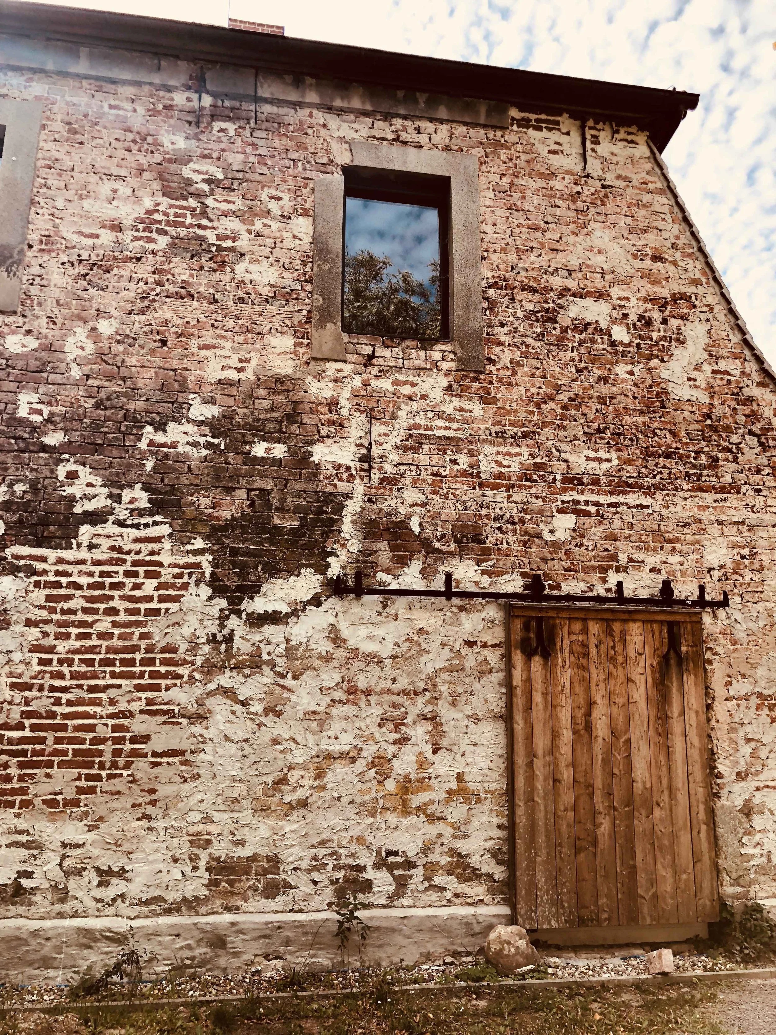 Nahaufnahme einer verwitterten Backsteinfassade am ehemaligen Brauhaus Briest mit rustikaler Holzschiebetür, Steinsockel und einem quadratischen Fenster im Dachgeschoss.