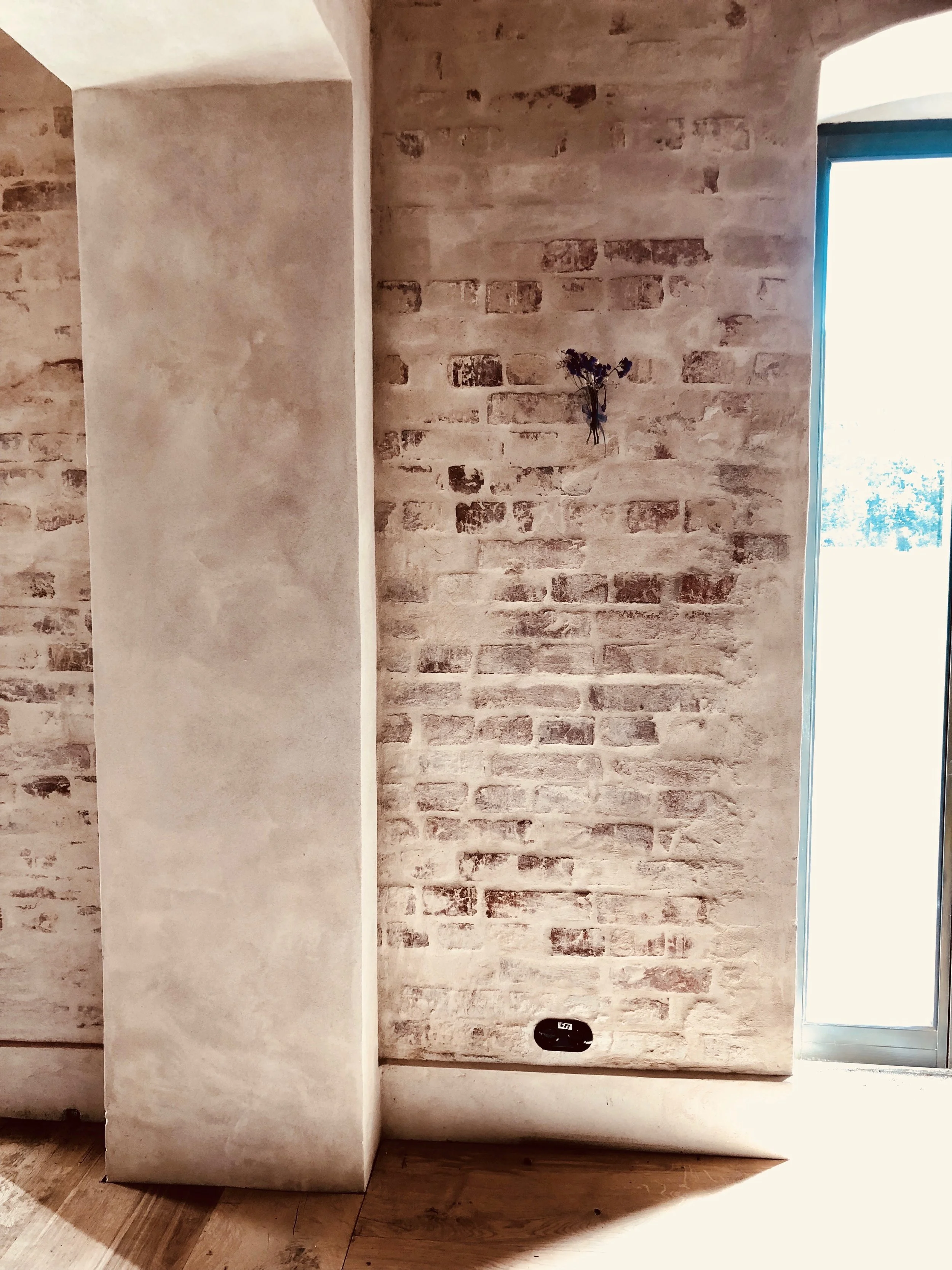 Detailaufnahme eines Innenraums mit einer glatt verputzten Betonsäule, einer angrenzenden Wand aus weiß geschlämmtem Backstein, einem Holzdielenboden und einer modernen Steckdosenunterdose. Im Hintergrund ist eine Glasfront sichtbar.