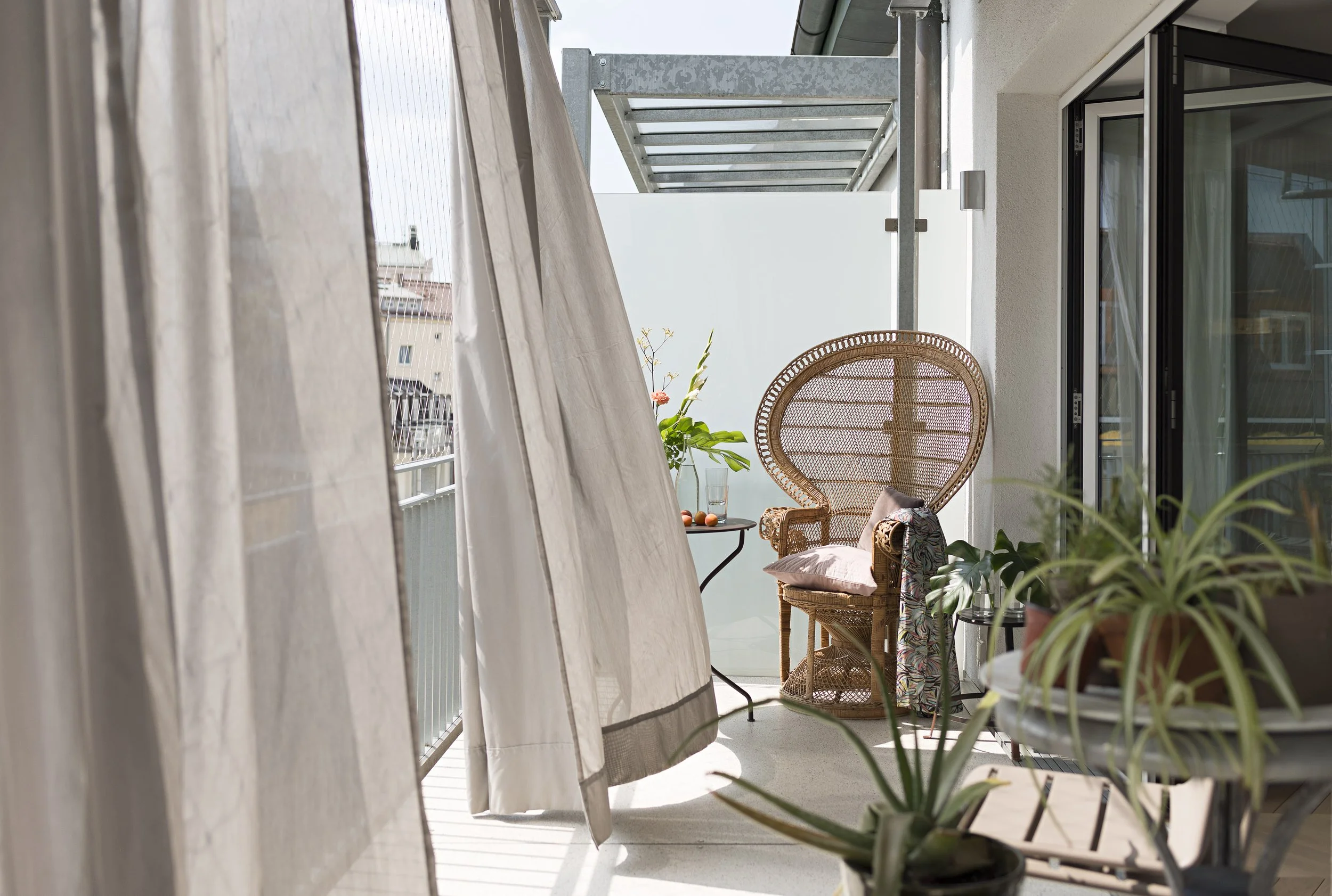 Hell erleuchteter Balkon mit einem großen Pfauensessel aus Rattan, Zimmerpflanzen und weichen Vorhängen, die im Wind wehen.