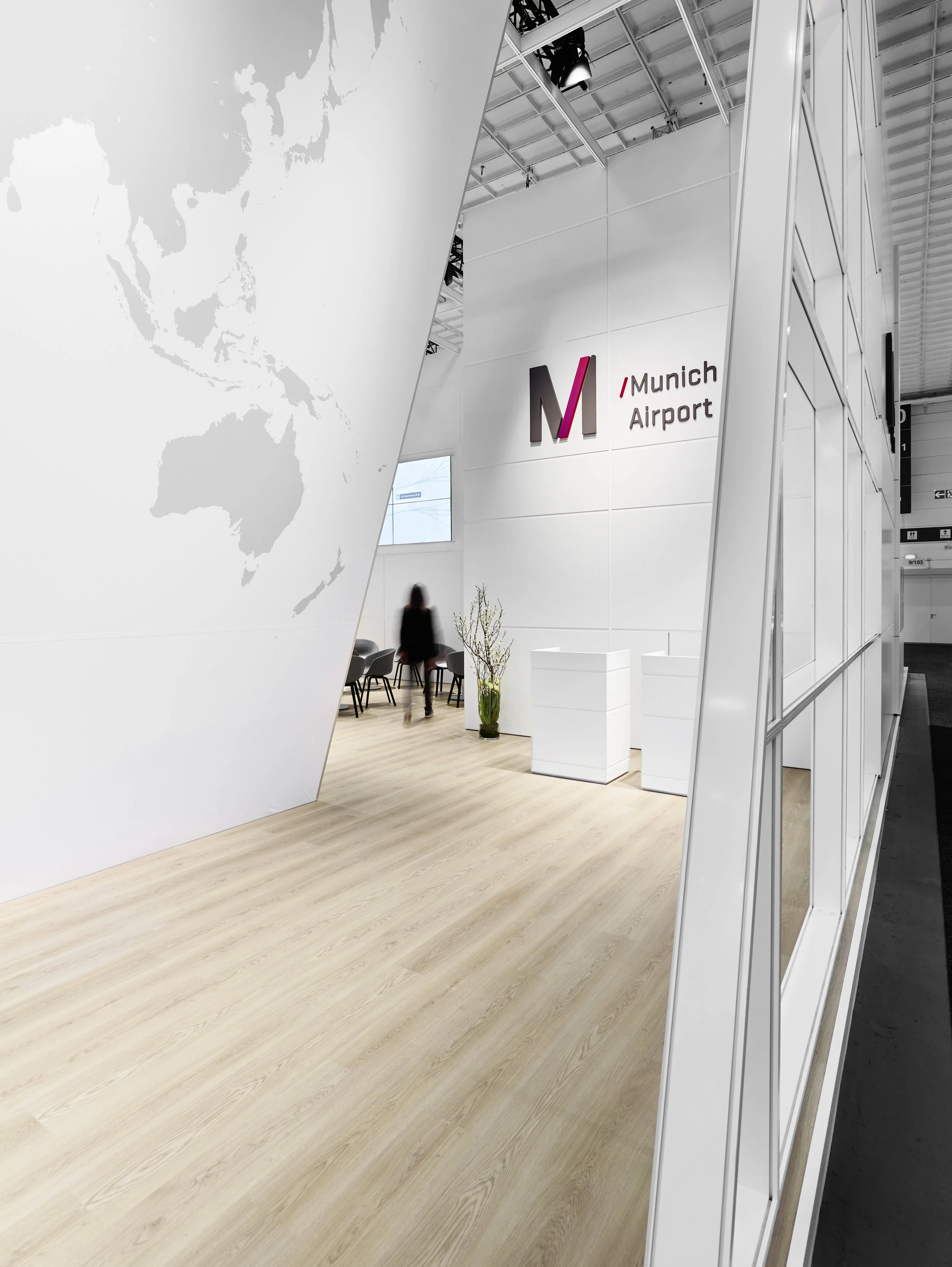 Hohe Wände des Messestands mit aufgedruckter Weltkarte, dem farbigen Logo „Munich Airport“, weißen Counter-Elementen und hellmoliertem Holzboden.