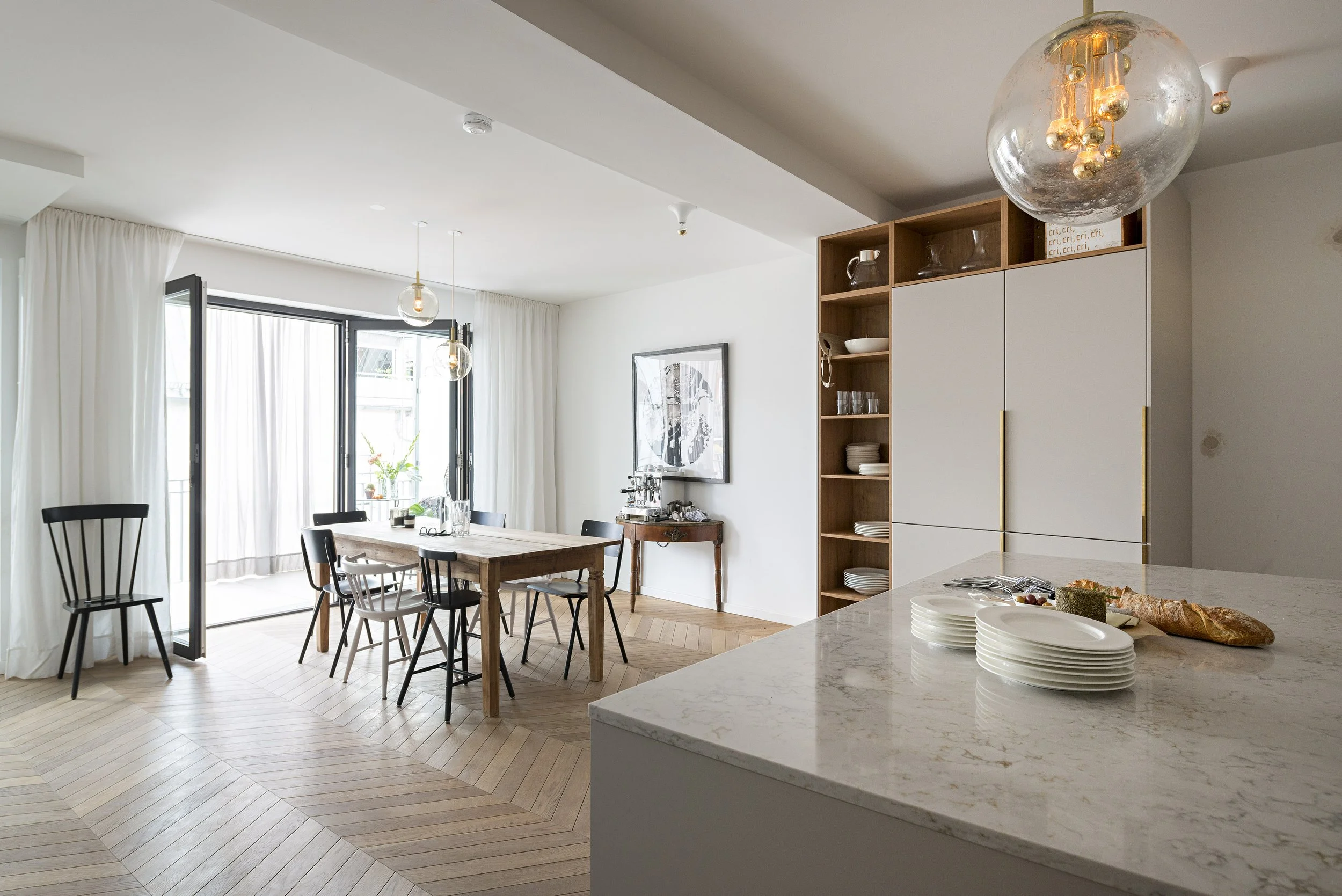Kücheninsel im Vordergrund mit Geschirr und Brot, dahinter ein Holztisch mit Stühlen und eine große Fensterfront mit Balkonzugang.