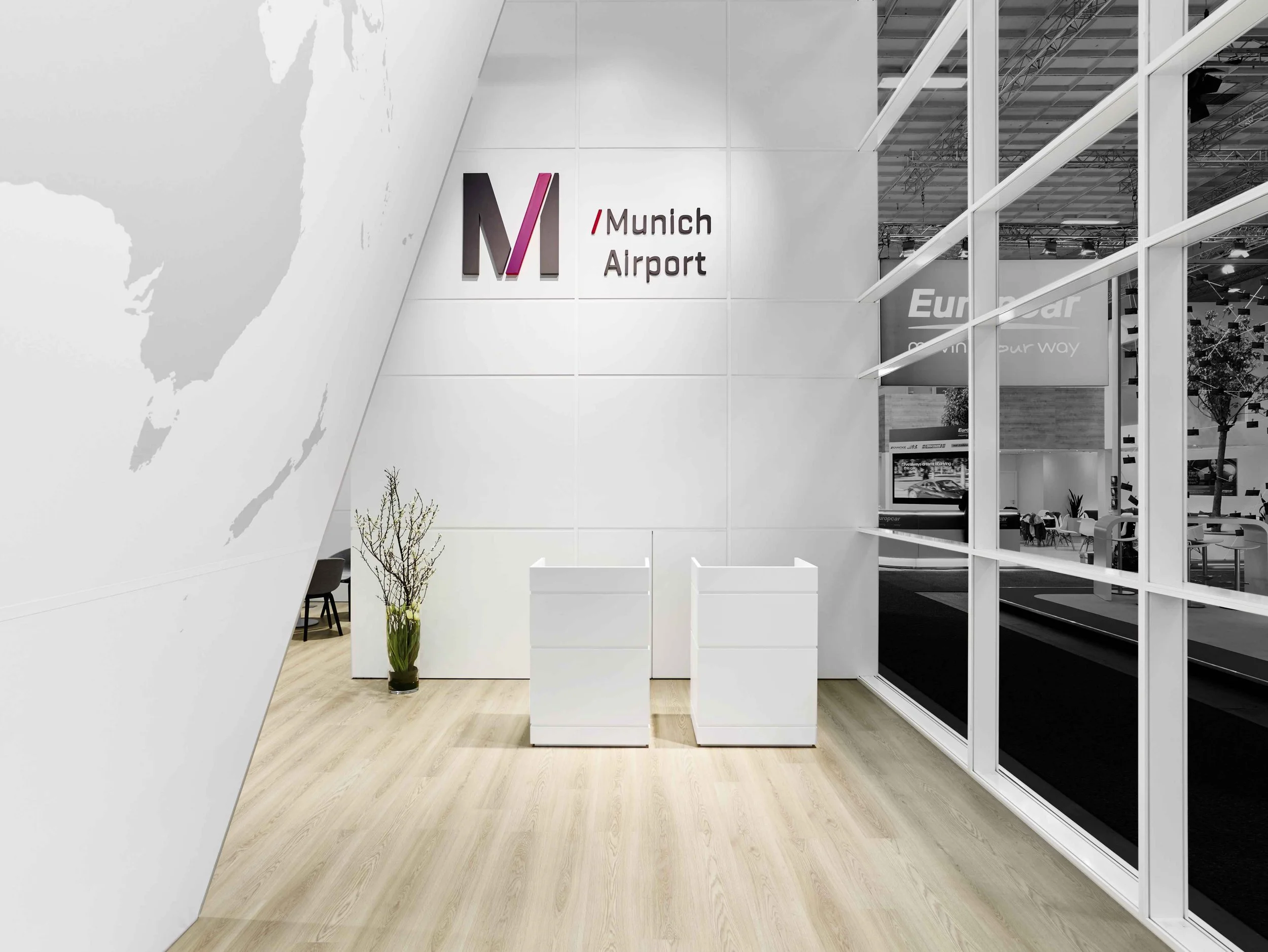 Moderner, heller Messestand des Flughafens München mit Holzboden, weißer Rückwand inklusive Logo „Munich Airport“, zwei minimalistischen weißen Empfangscountern und einer Glasfront.