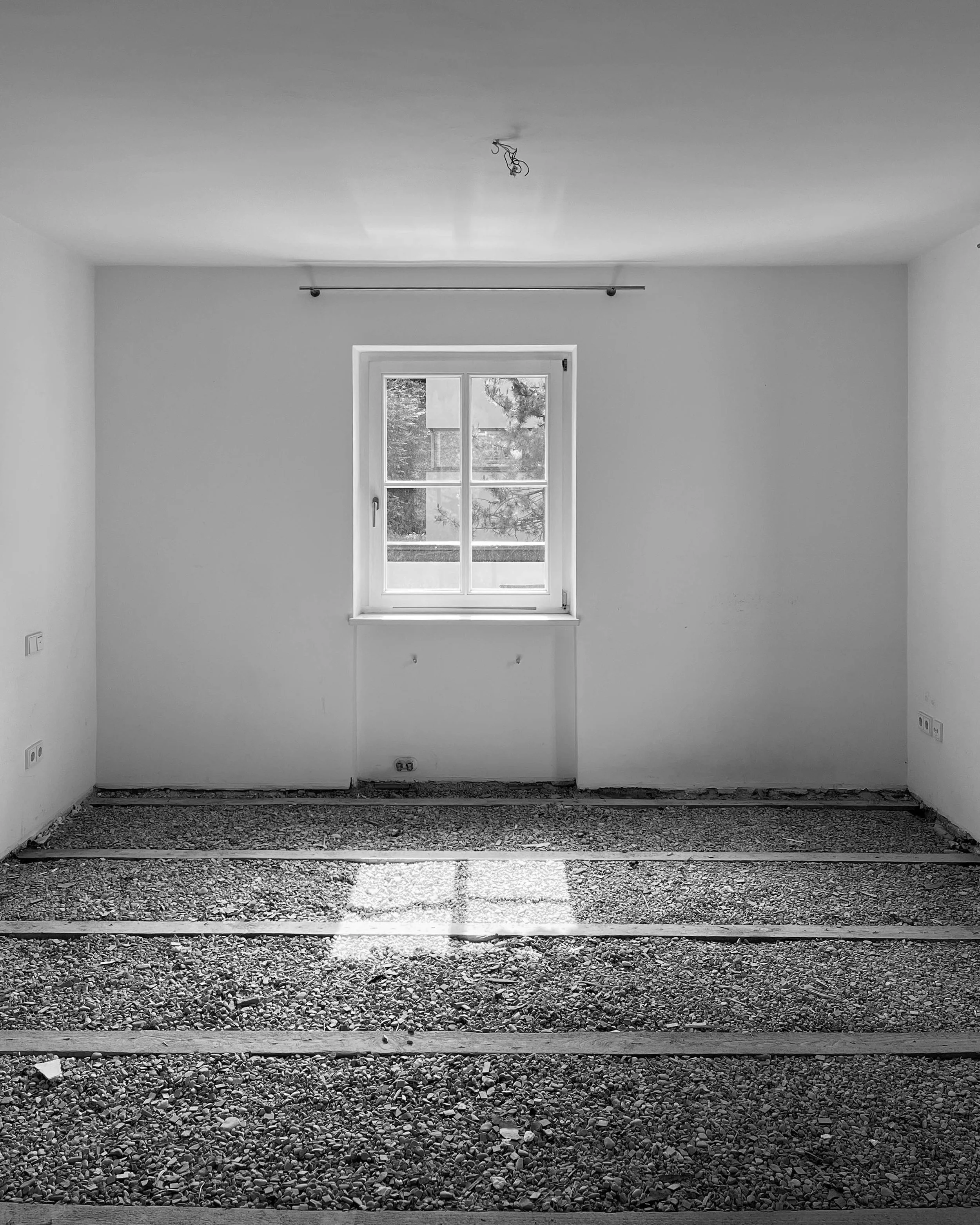 Aufnahme eines Innenraums während der Sanierung mit freigelegter Fehlboden-Schüttung zwischen den Holzbalken, weiß verputzten Wänden und einem zentralen Sprossenfenster.