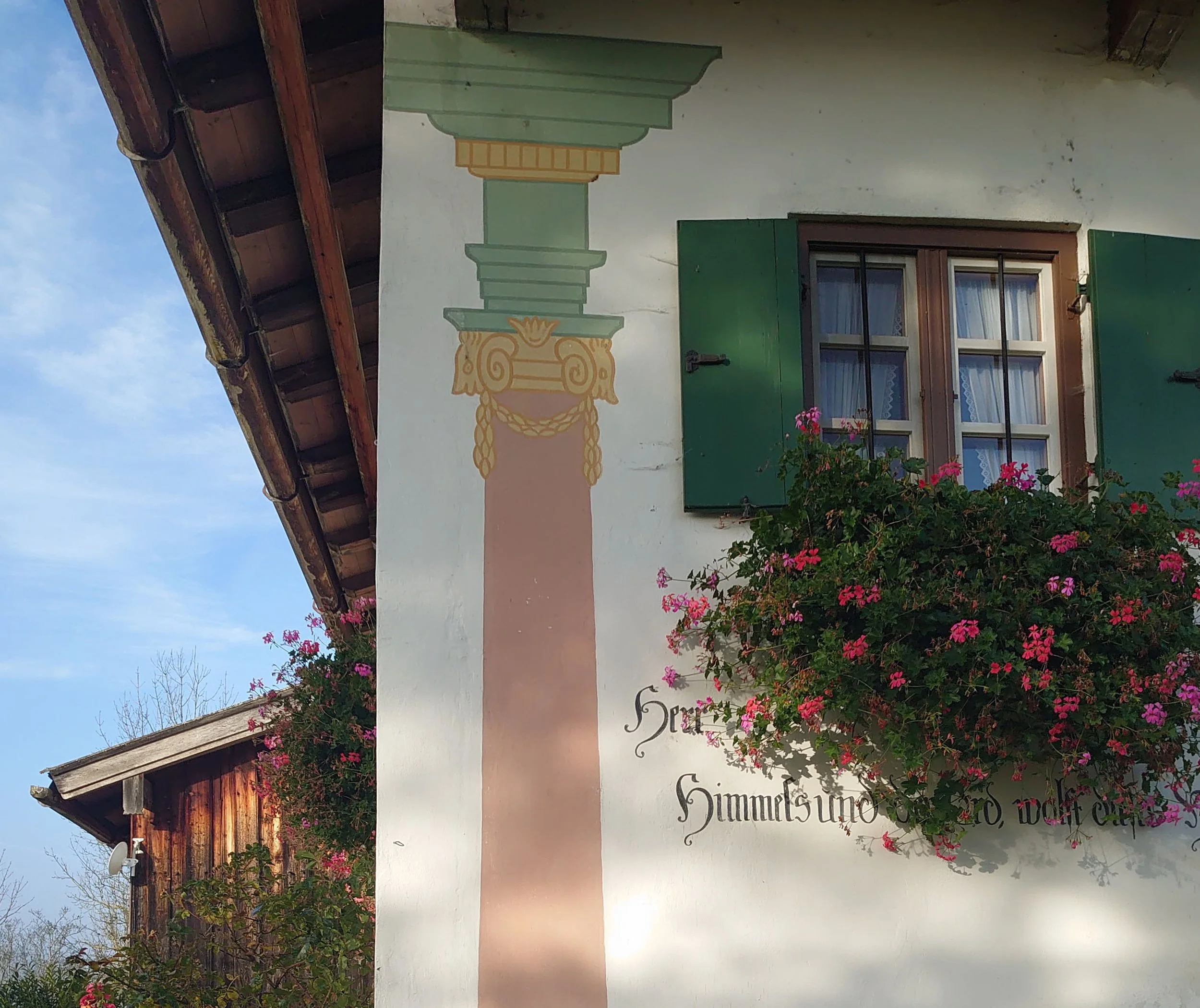 Nahaufnahme einer sanierten Altbau-Fassade mit traditioneller Lüftlmalerei, grünen Klappläden, blühenden Geranien im Blumenkasten und altdeutscher Frakturschrift.