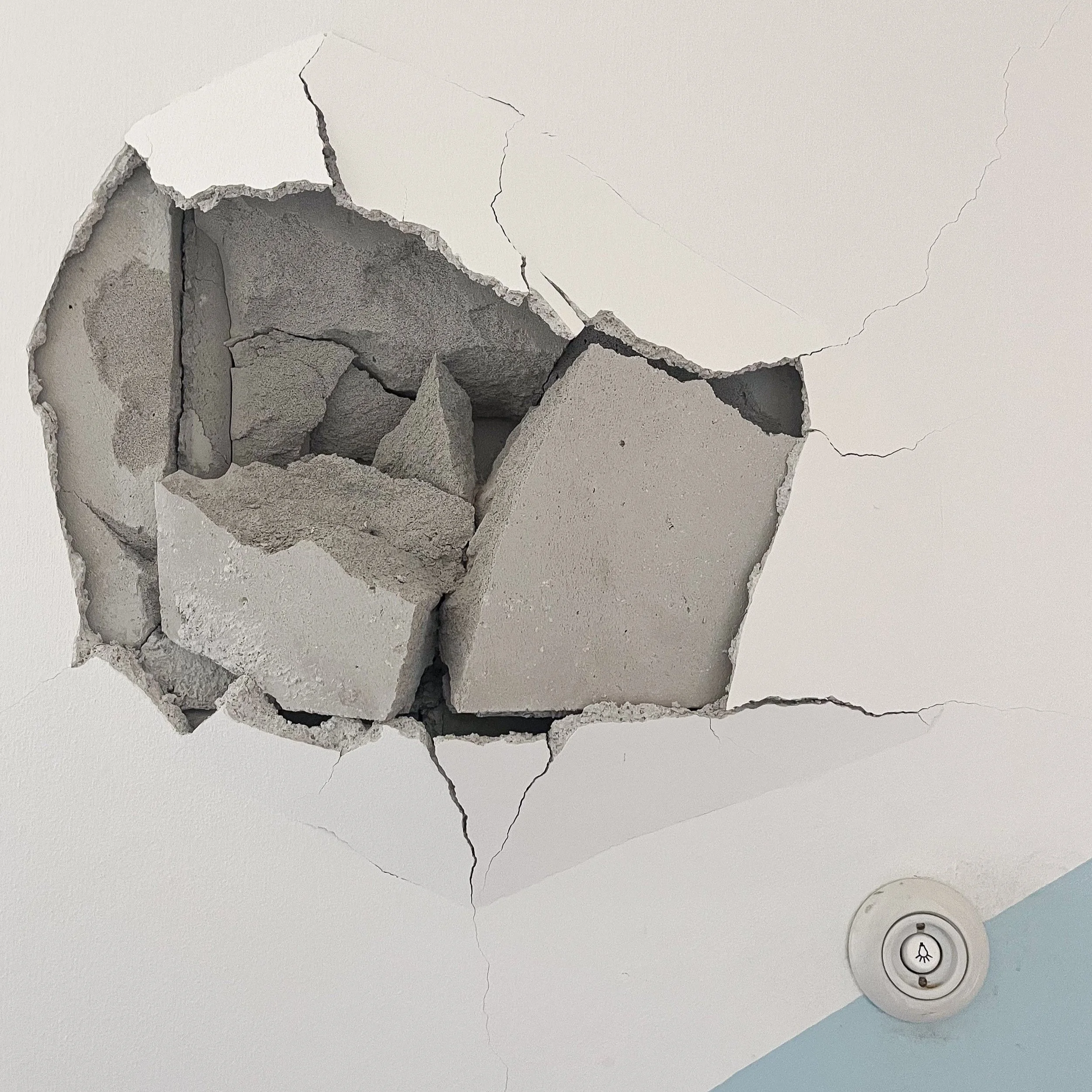 Beschädigte Innenwand mit großflächigem Putzabplatzer, sichtbarem Mauerwerk und mehreren Rissen.