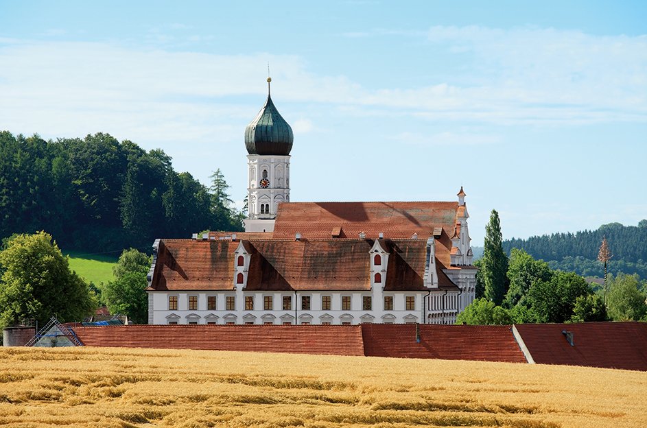 Panorama-Außenaufnahme einer historischen Schlossanlage mit Zwiebelturm, barocken Renaissance-Giebeln, rotem Ziegeldach und Schlosskirche, eingebettet in Hügellandschaft hinter einem Getreidefeld.