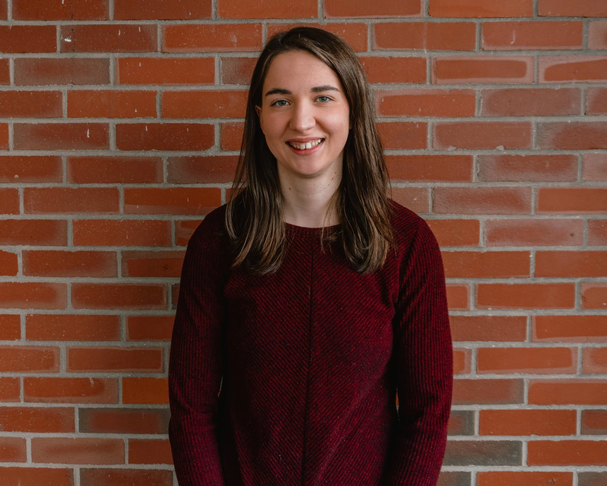 A woman with shoulder-length brown hair, wearing a green top, smiling with visible teeth against a gray mottled background.