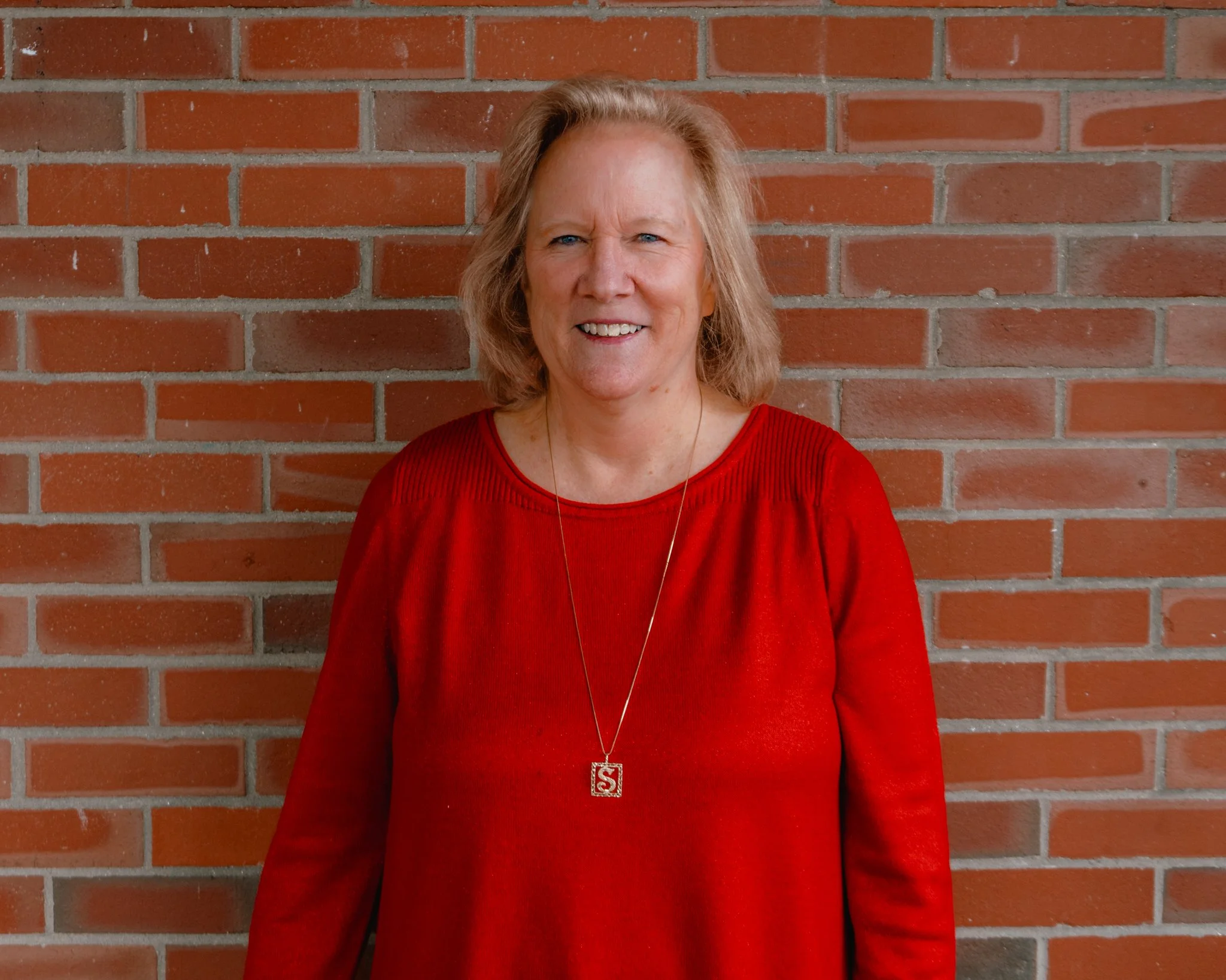 Older woman with blonde hair smiling, wearing a black top and a patterned blouse with a bow tie at the collar, against a gray background.