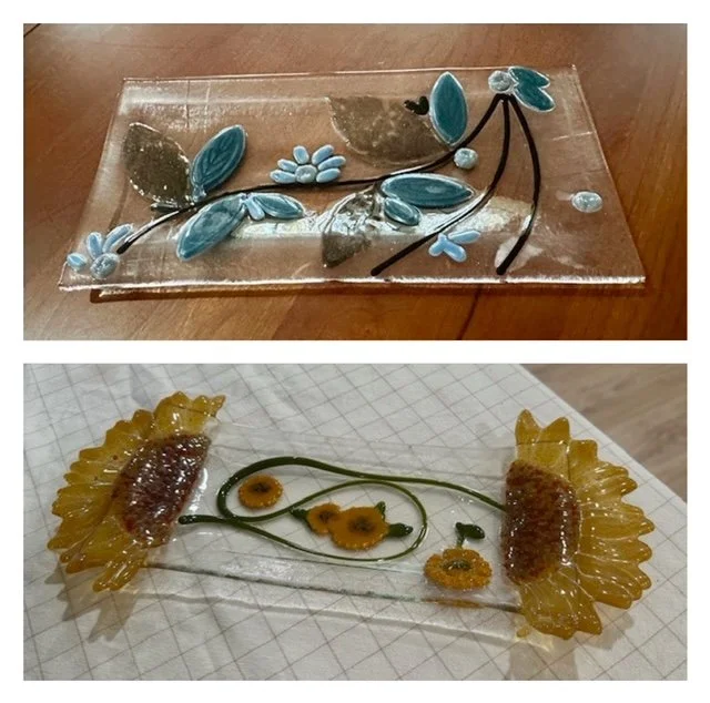 Two fused glass art pieces, one featuring blue leaves and flowers on a clear background, and the other resembling yellow and orange flowers with green stems on a light background.