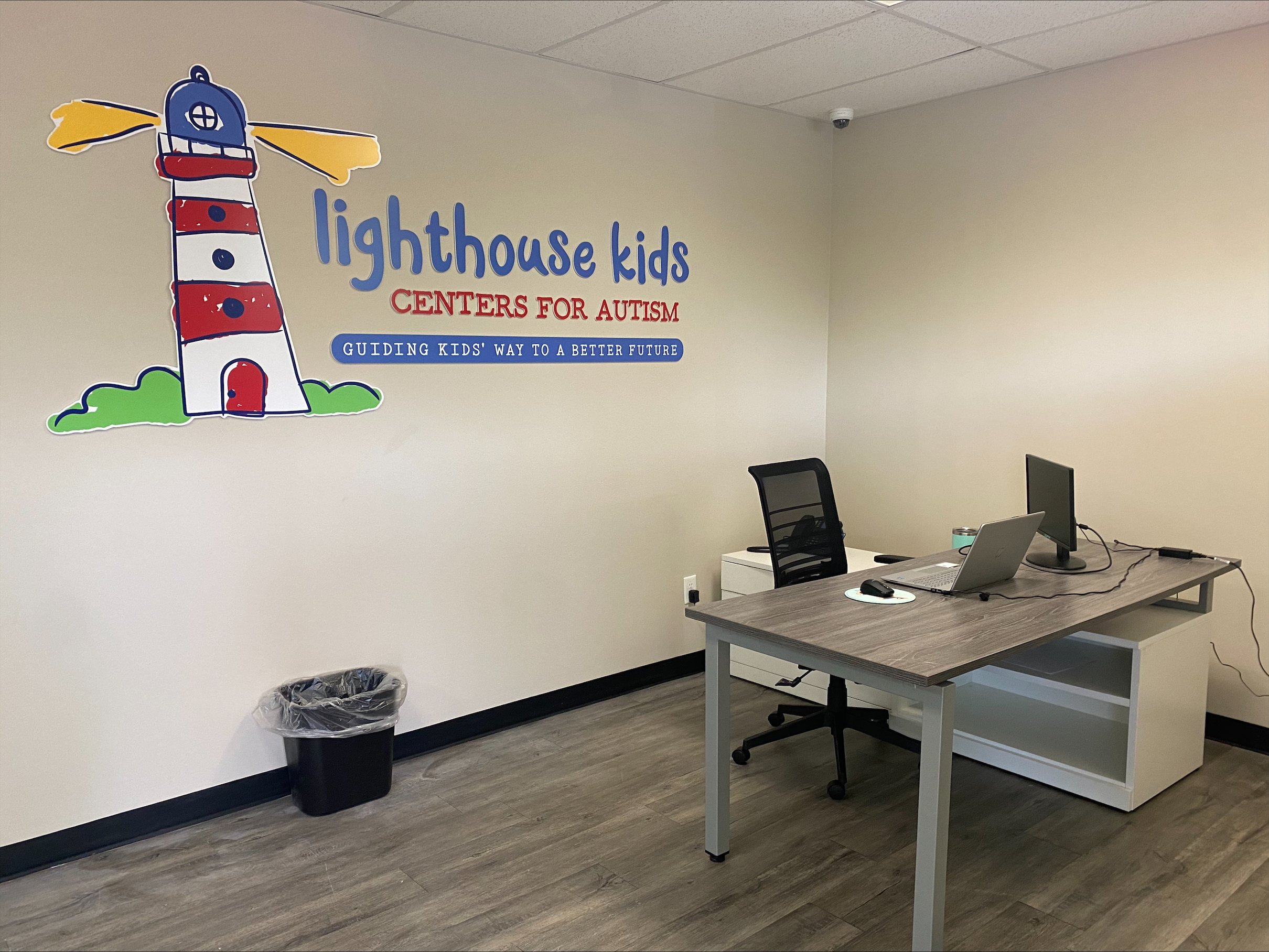 Office interior with "Lighthouse Kids Centers for Autism" wall decal, desk with laptop and monitor, chair, and trash bin.