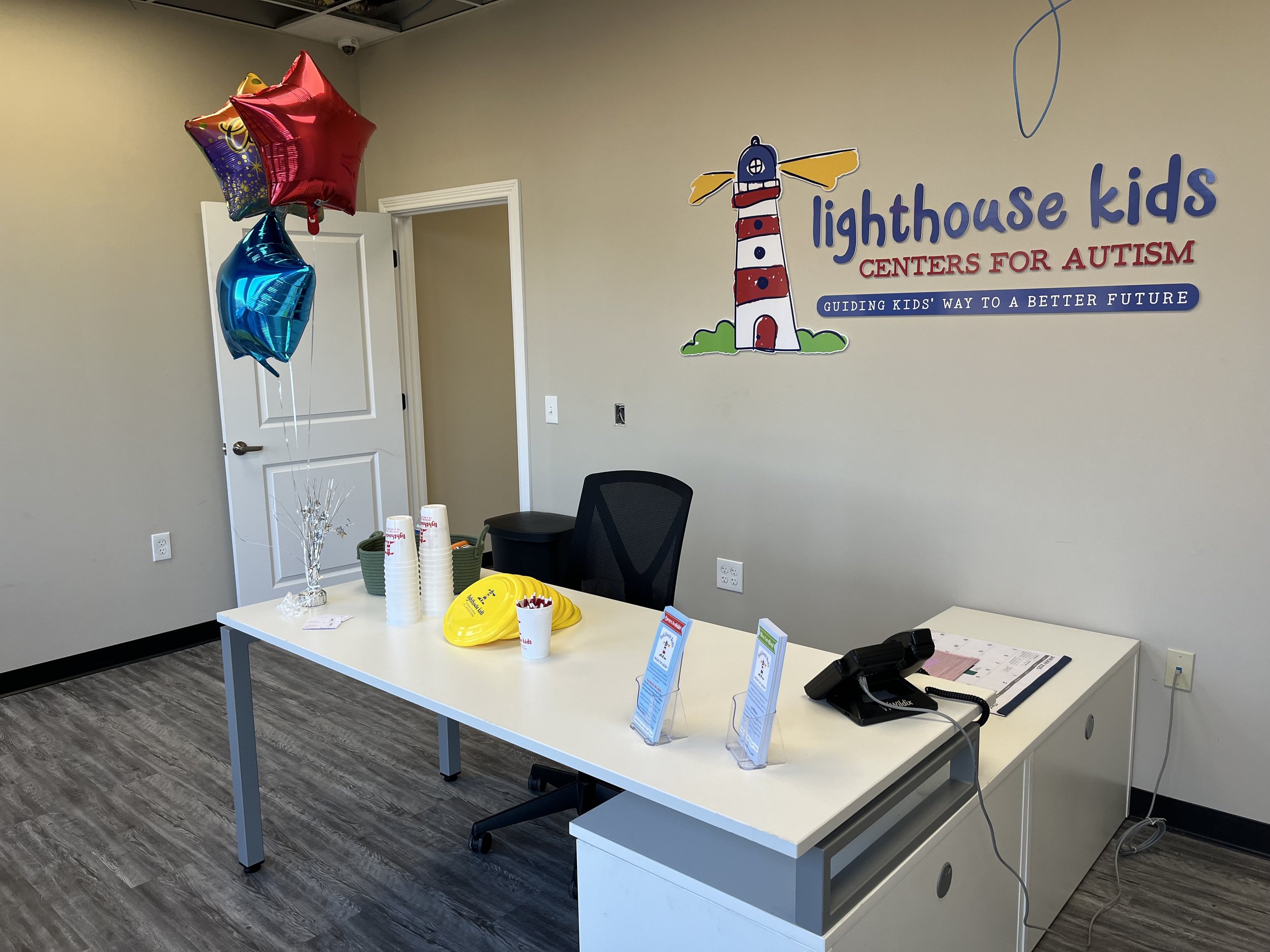 Office reception area with balloons, cups, brochures, and the logo "lighthouse kids centers for autism" on the wall.