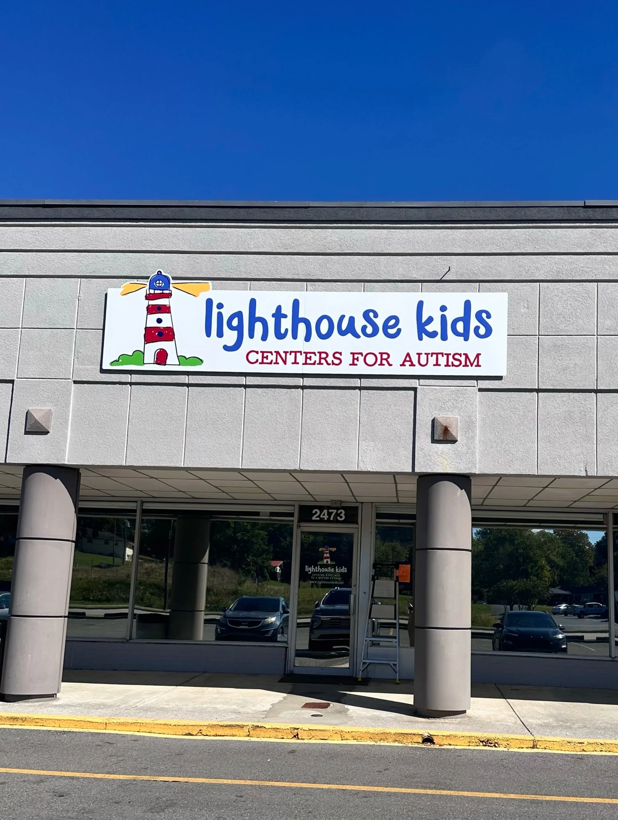 Exterior view of a building with a sign for Lighthouse Kids Centers for Autism, featuring a cartoon lighthouse logo, during daytime with clear sky.