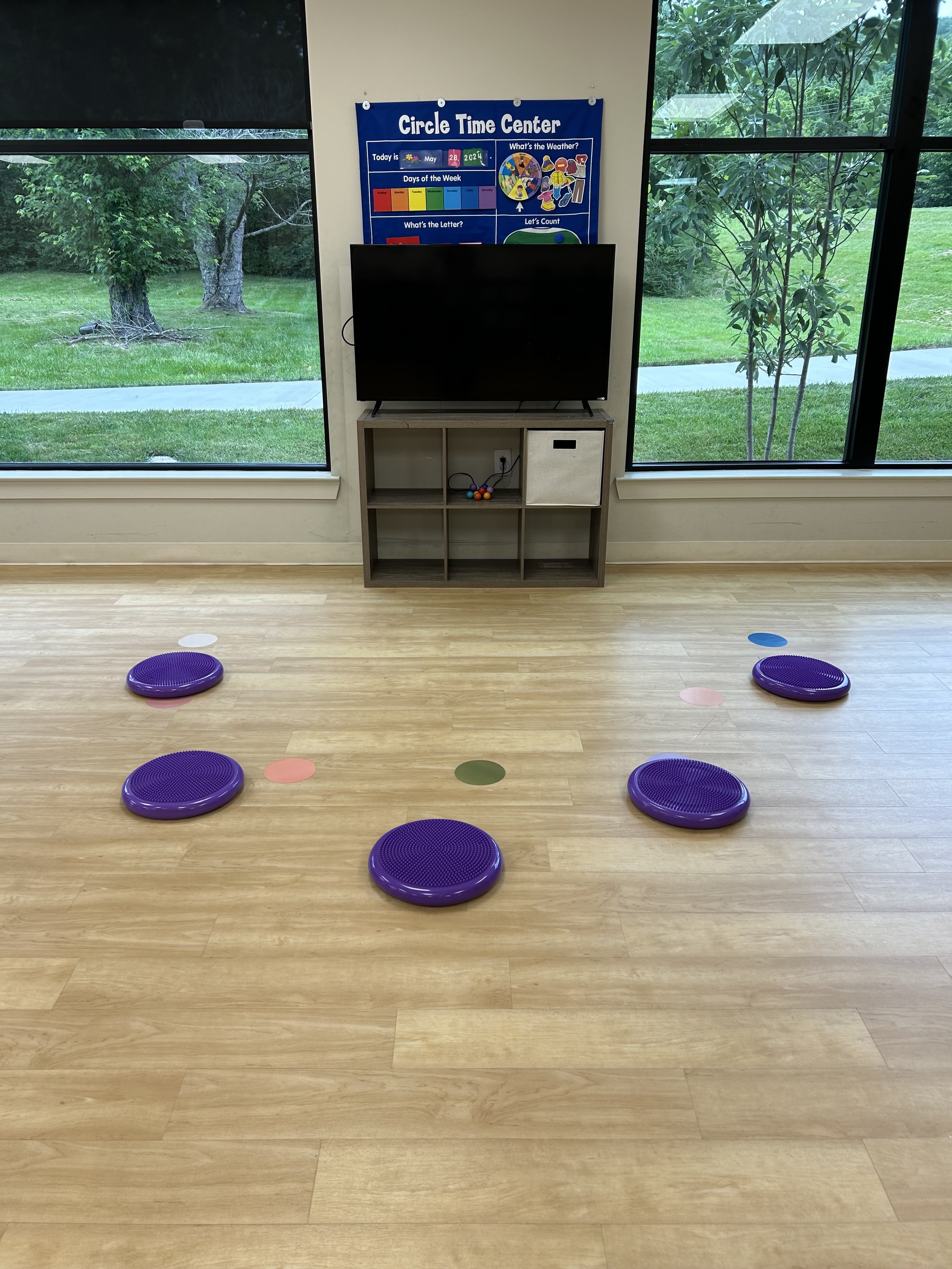 Preschool classroom with activity setup, purple cushion seats, circle time poster, TV on a shelf, large windows, light wood flooring.