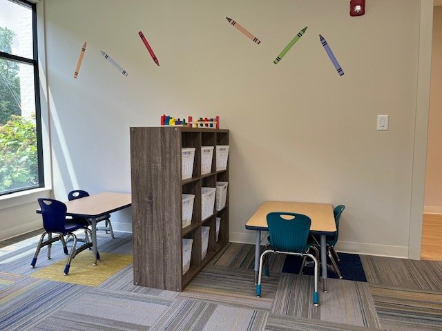 Classroom corner with two small tables and chairs, a shelf with bins, colorful block toys, and crayon decals on the wall.