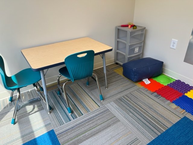 Classroom corner with table, two blue chairs, shelf, cushion, and colorful textured tiles on the floor.