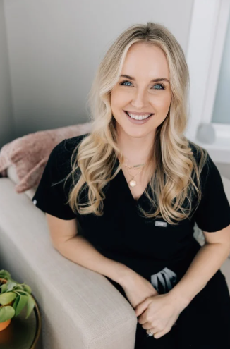 A smiling woman with blonde hair, wearing black gloves and jewelry, holding a small syringe filled with purple liquid in her right hand against a plain white background.