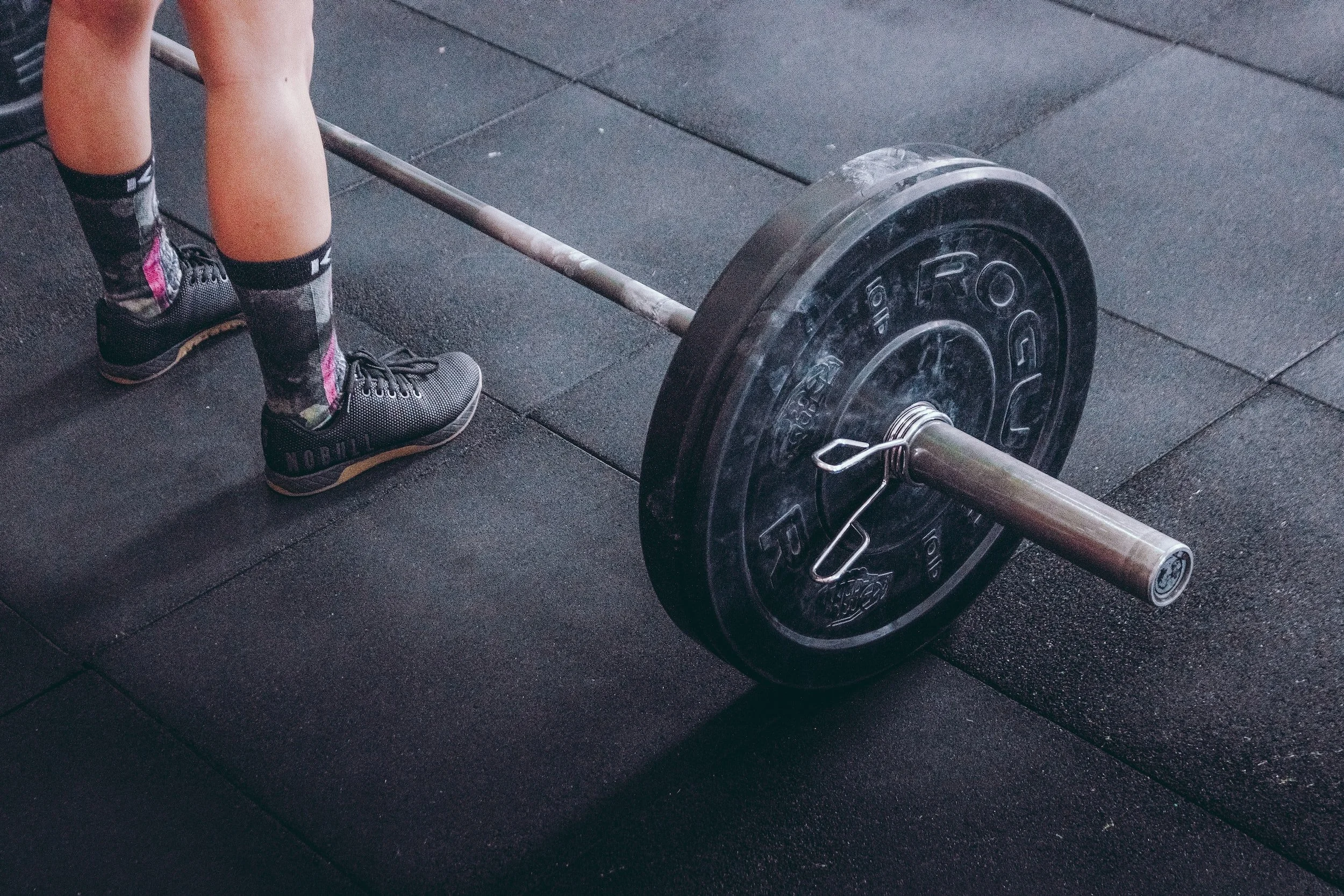Person standing beside a loaded barbell on a gym floor, wearing black shoes and socks with a floral pattern.