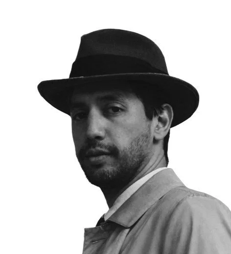 Black and white portrait of a man wearing a fedora hat and a jacket, looking over his shoulder.