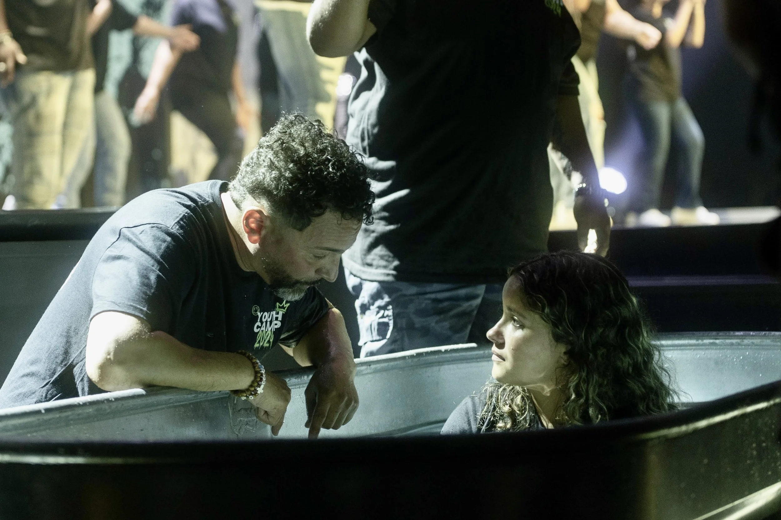 A man and a young person at a baptism event, with the man leaning over a metal baptismal pool as the young person looks at him. Other people are in the background.