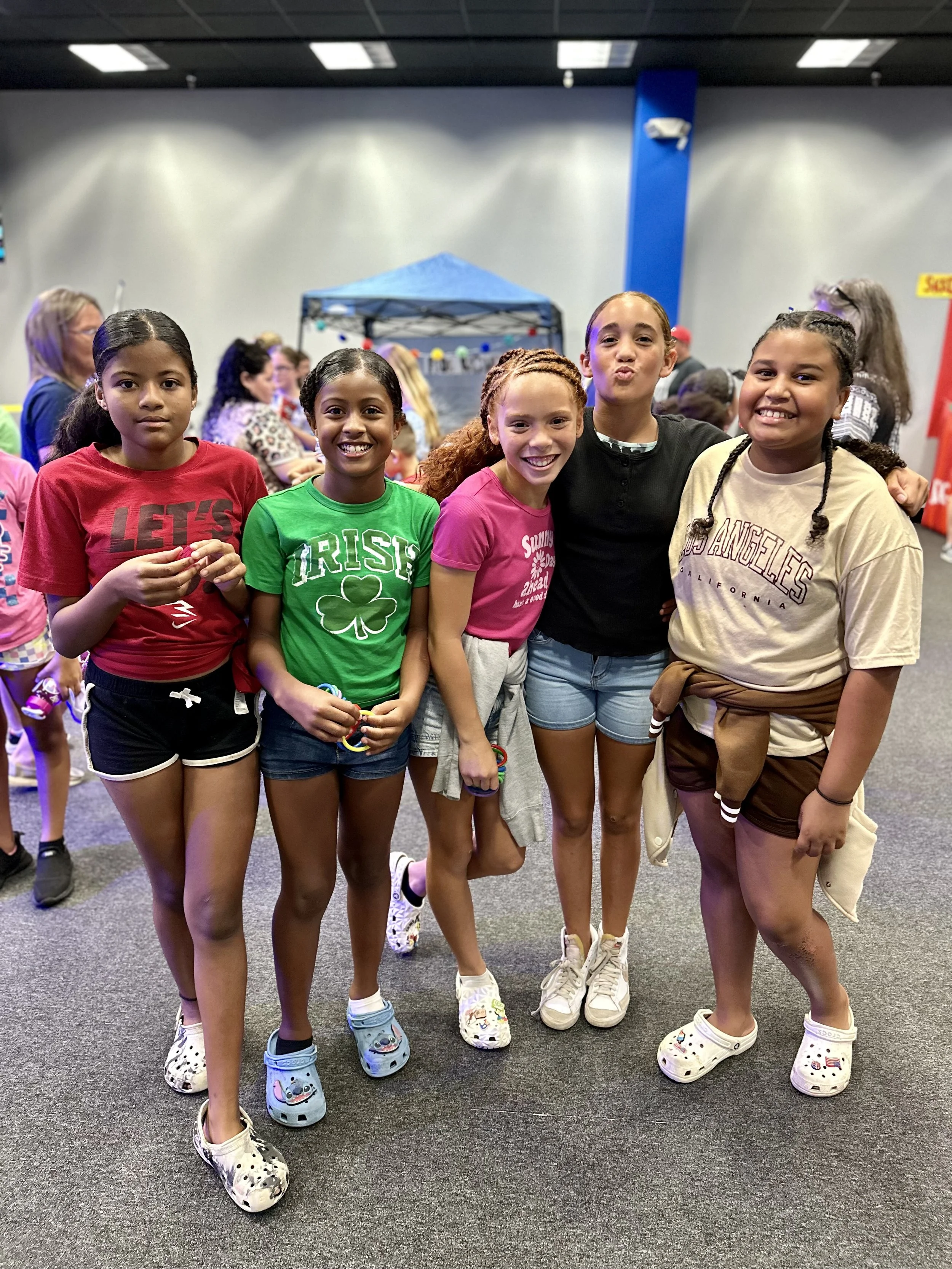 Group of five girls smiling indoors, wearing casual clothing and Crocs.