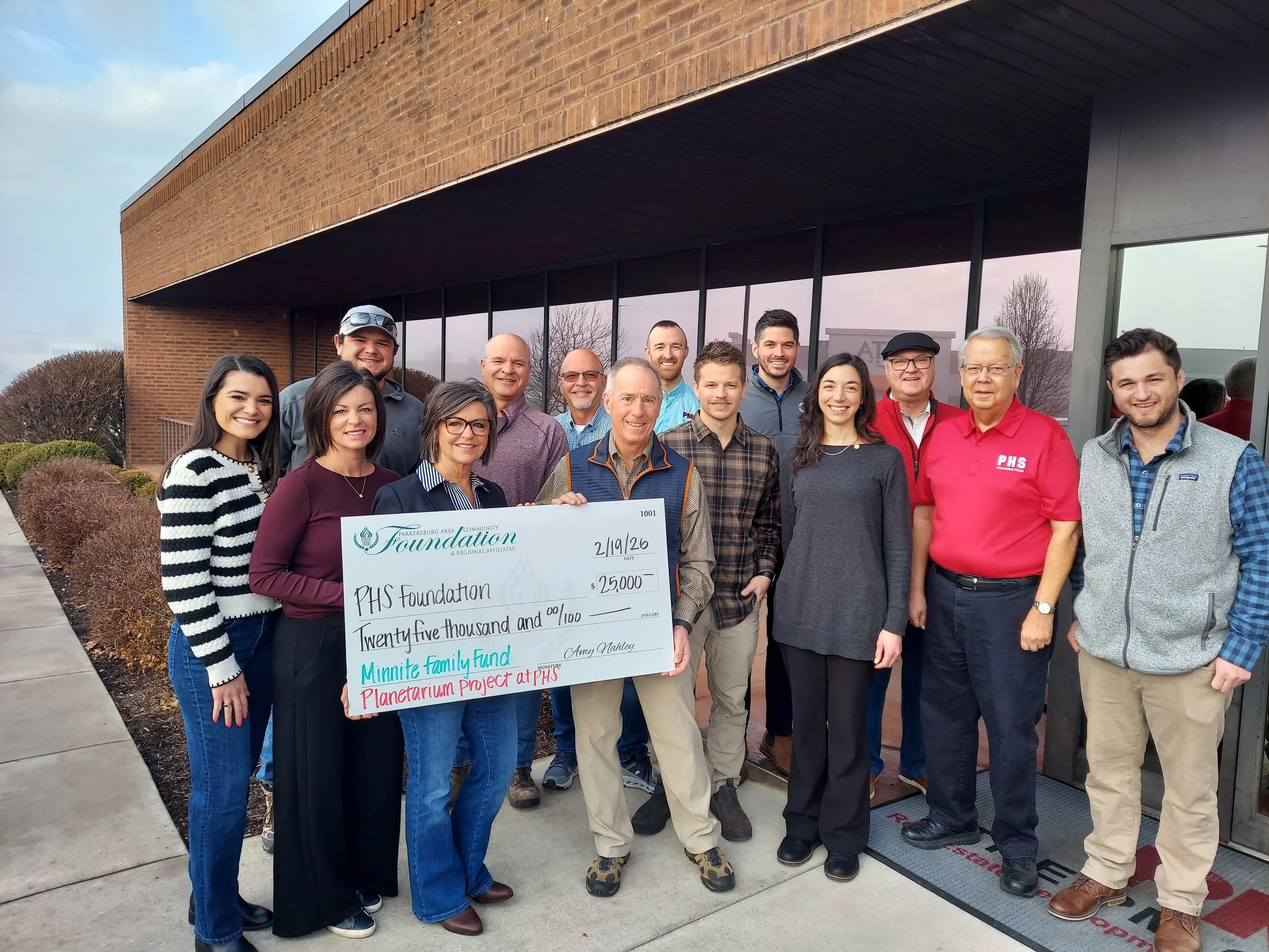 Group of people standing outside a building, smiling, holding a large check for $25,000 made out to PHS Foundation, during a fundraising event.
