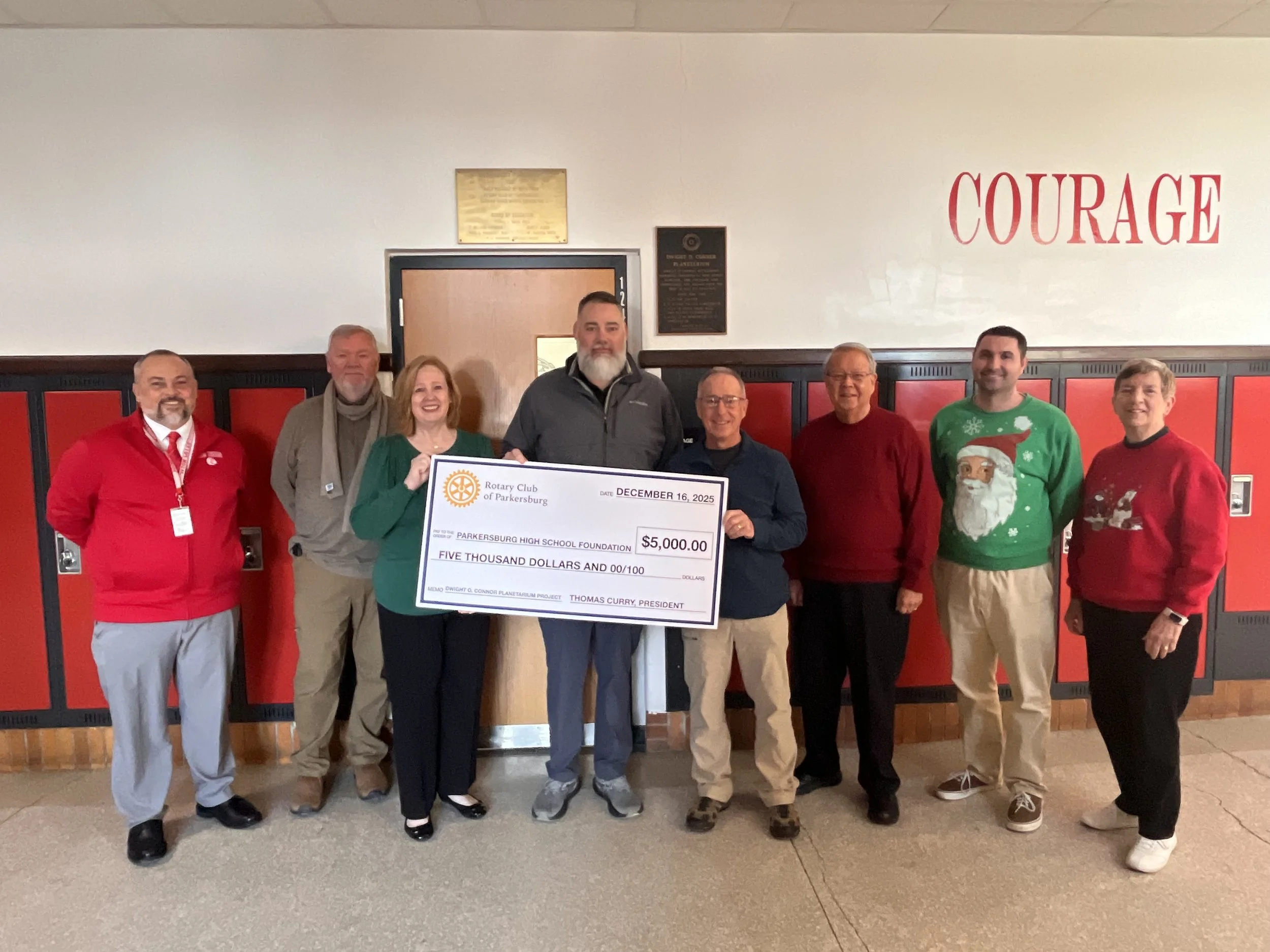 A group of ten people standing indoors in front of red and black lockers, holding a large check for $5,000 made out to the Parkersburg High School Foundation, dated December 16, 2025, with some individuals wearing holiday sweaters and others dressed in casual business attire.