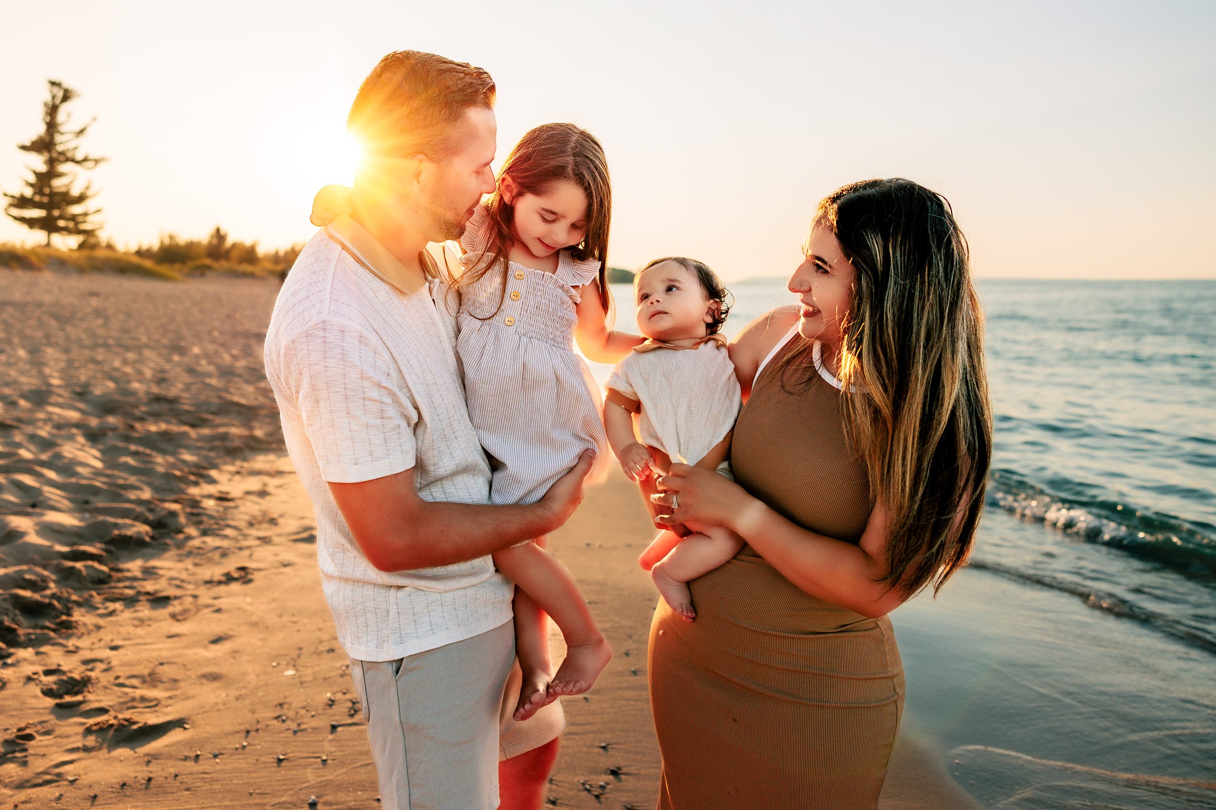 A family of four enjoying a moment on the beach at sunset, with the parents holding their two young children, all smiling and looking at each other.