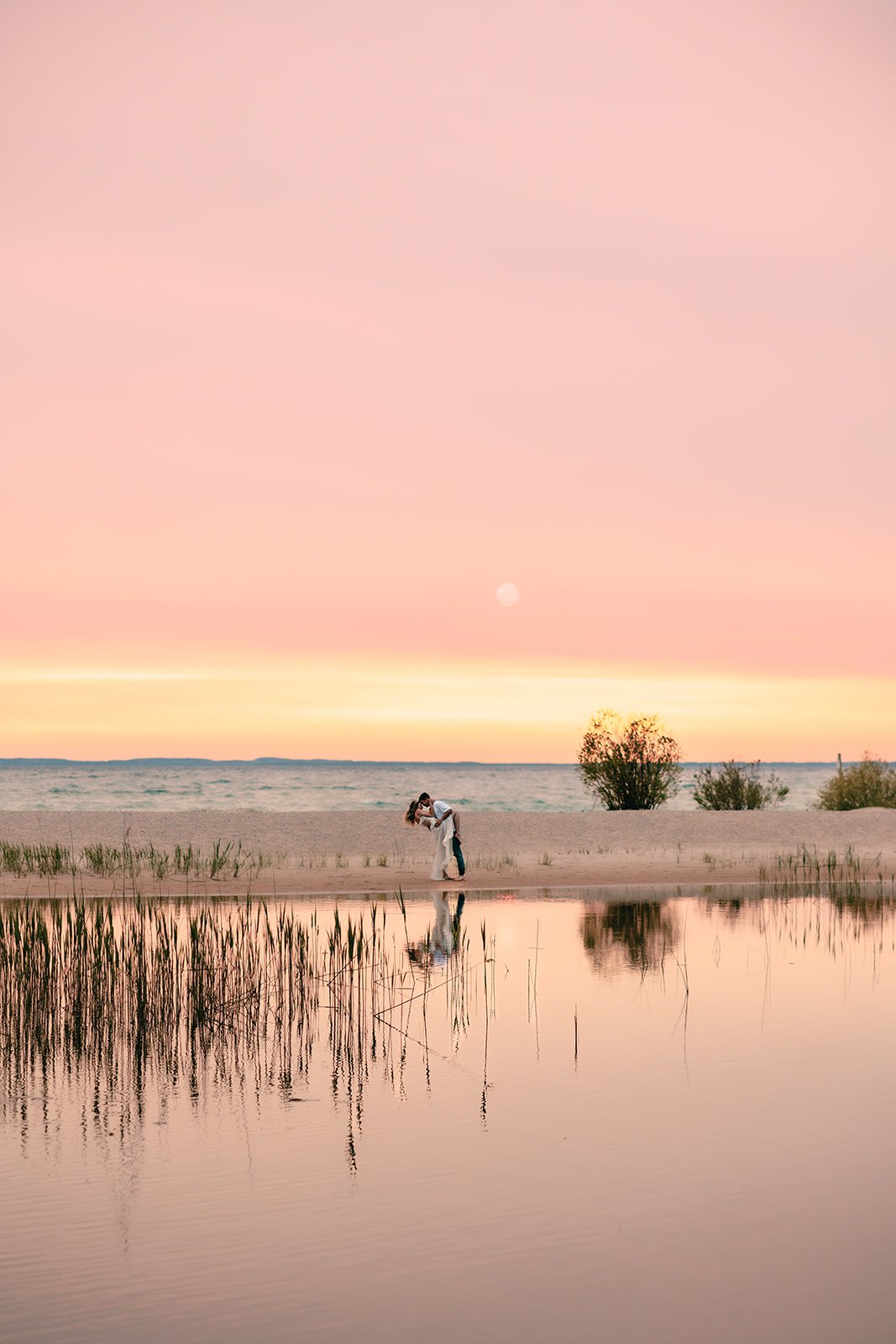 Couple kissing on the beach during sunset, with pink sky, water reflection, and trees in the background.