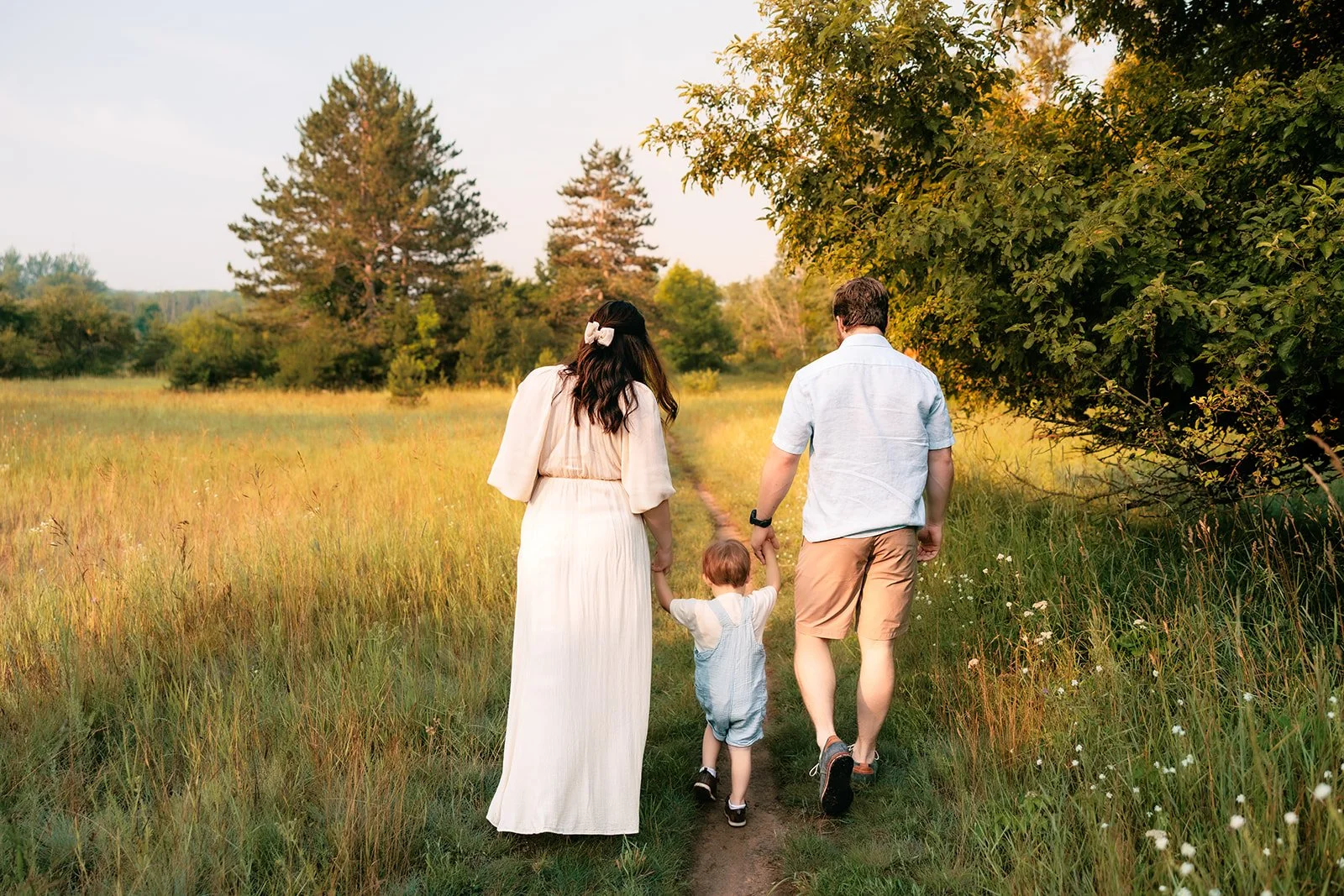A family of three walking hand in hand along a nature trail through a grassy field with trees at sunset in traverse city