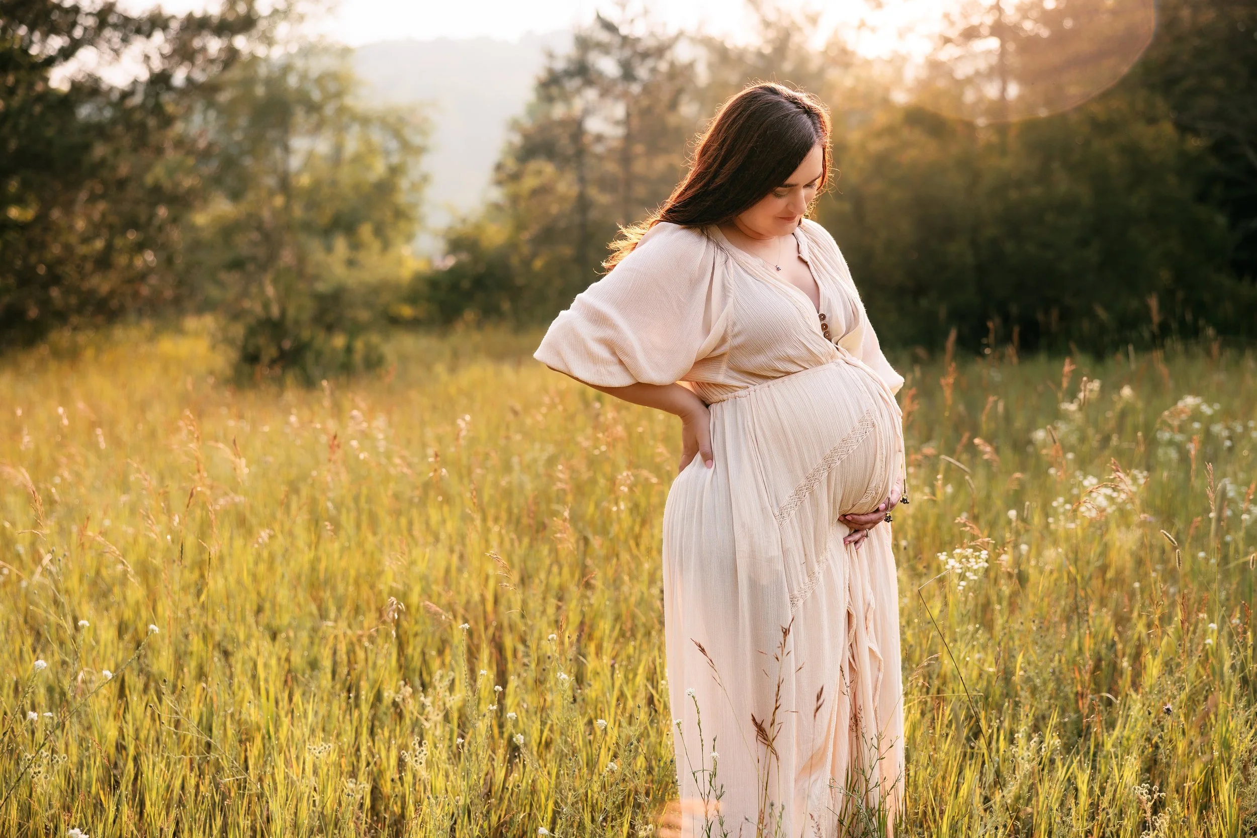 A pregnant woman standing in a grassy field during sunset, gently holding her belly with one hand and looking down.