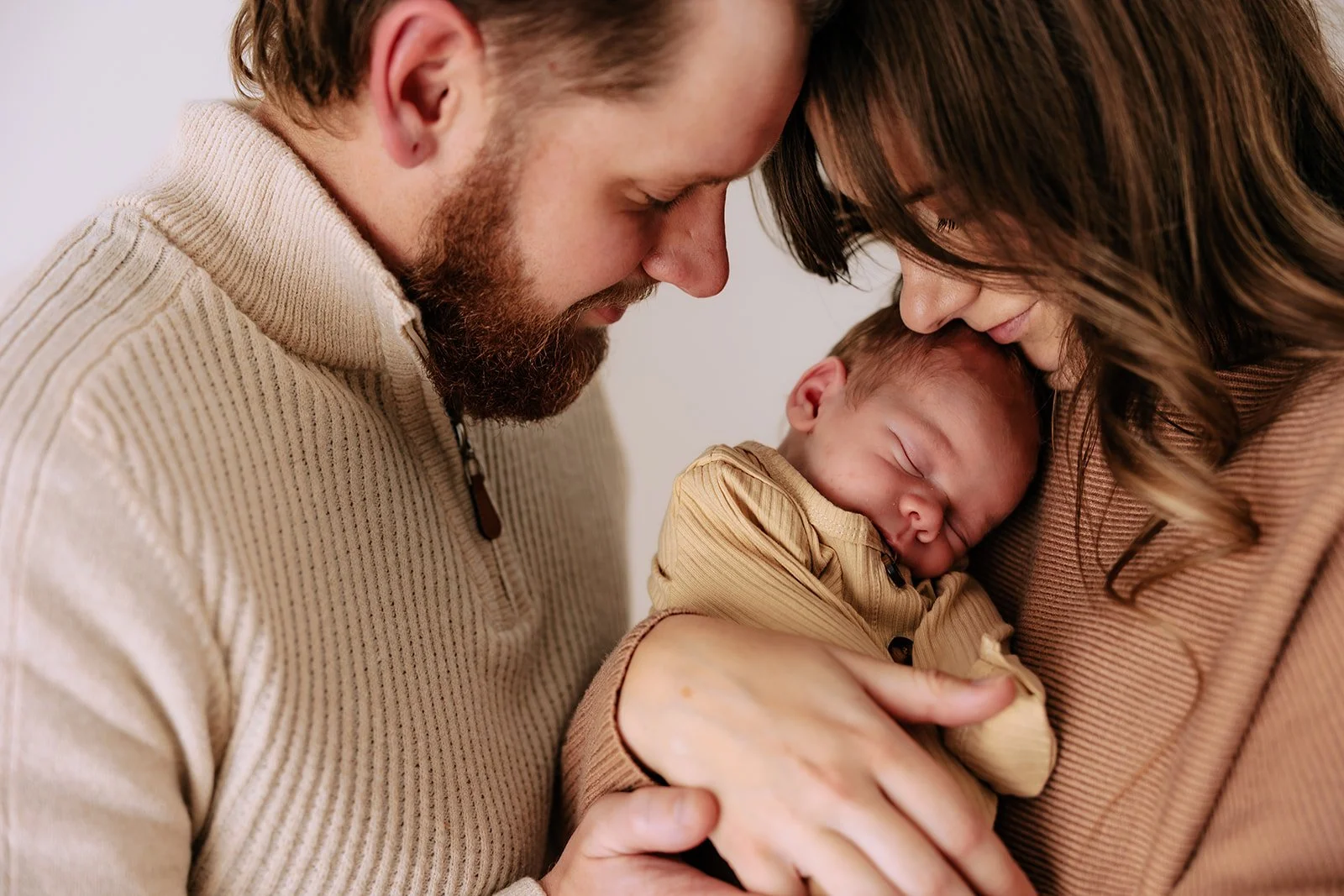 A family of three, a mother, father, and newborn baby, sharing a tender moment with their foreheads touching and the baby's face nestled between the parents' faces.