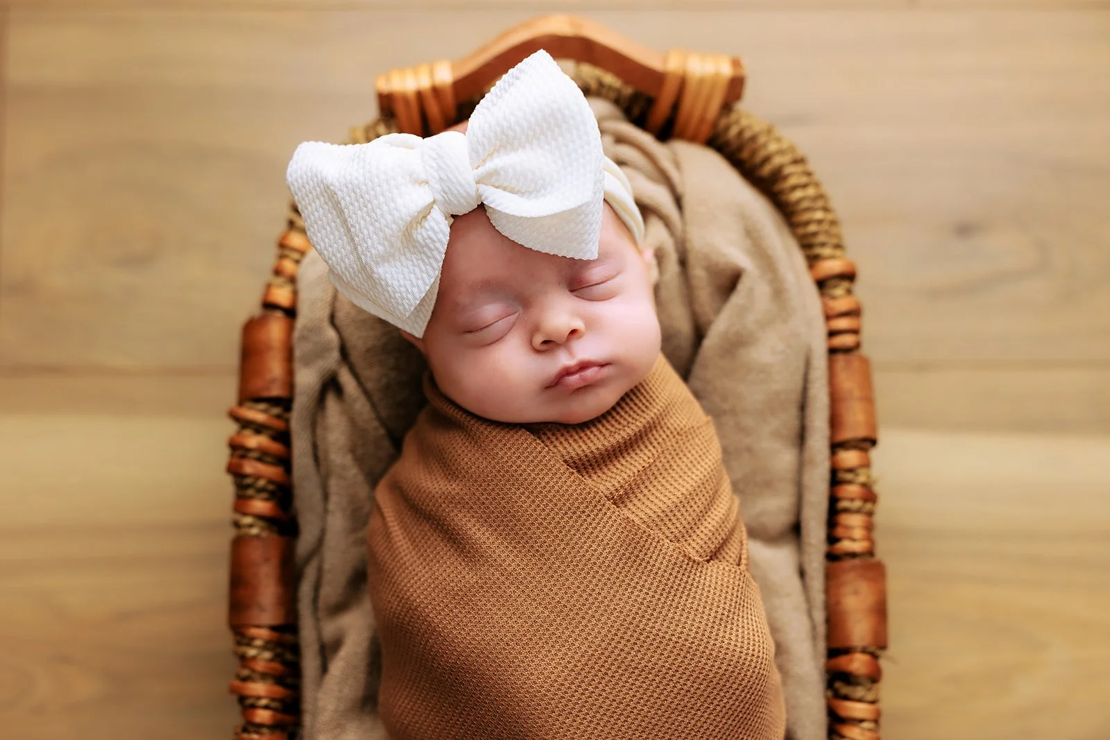 A sleeping baby wrapped in a brown blanket, lying in a wicker basket with a large white bow on their head in northern michigan