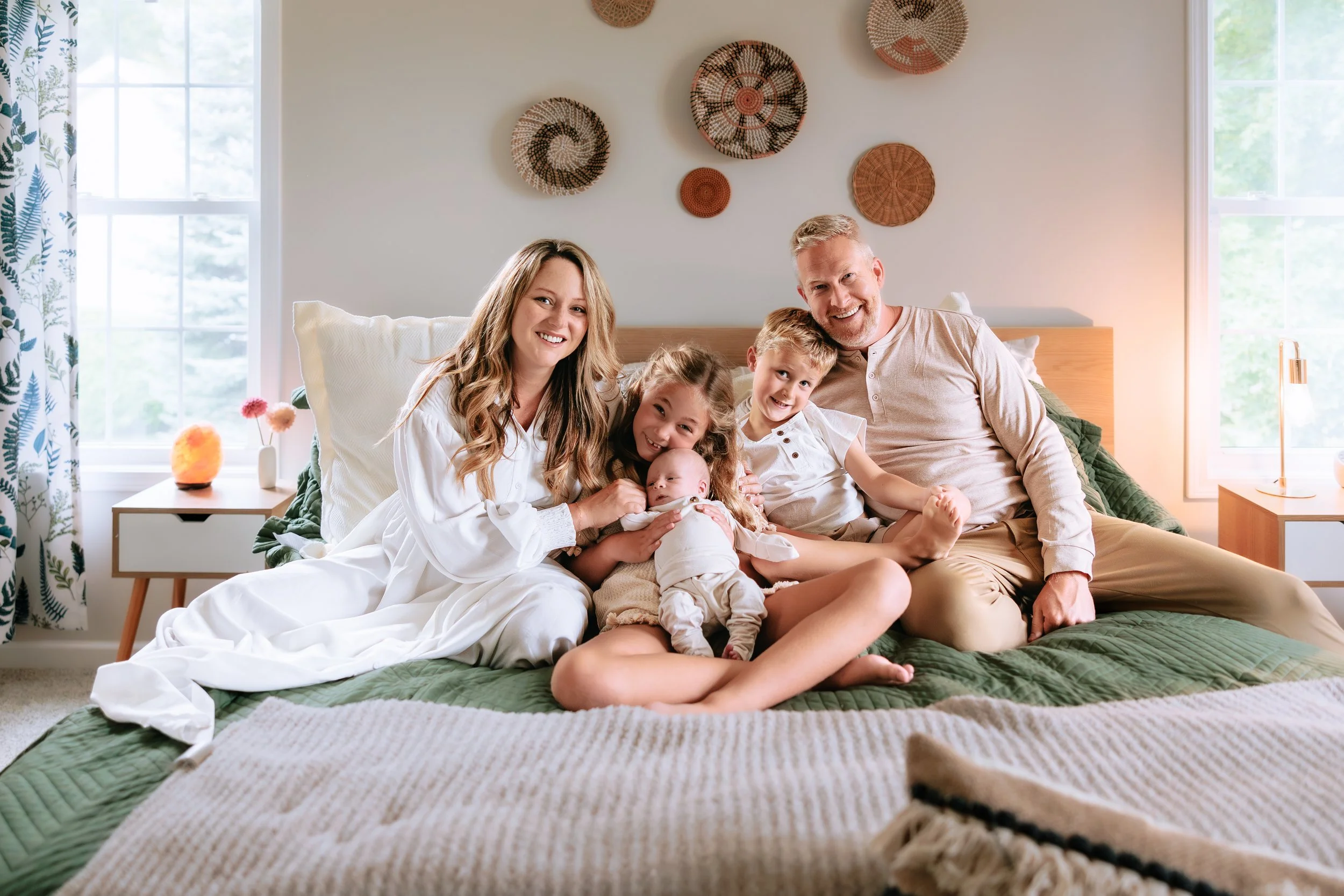 A happy family sitting on a bed in a bright bedroom, smiling at the camera. The family includes two parents, two children, and a baby.