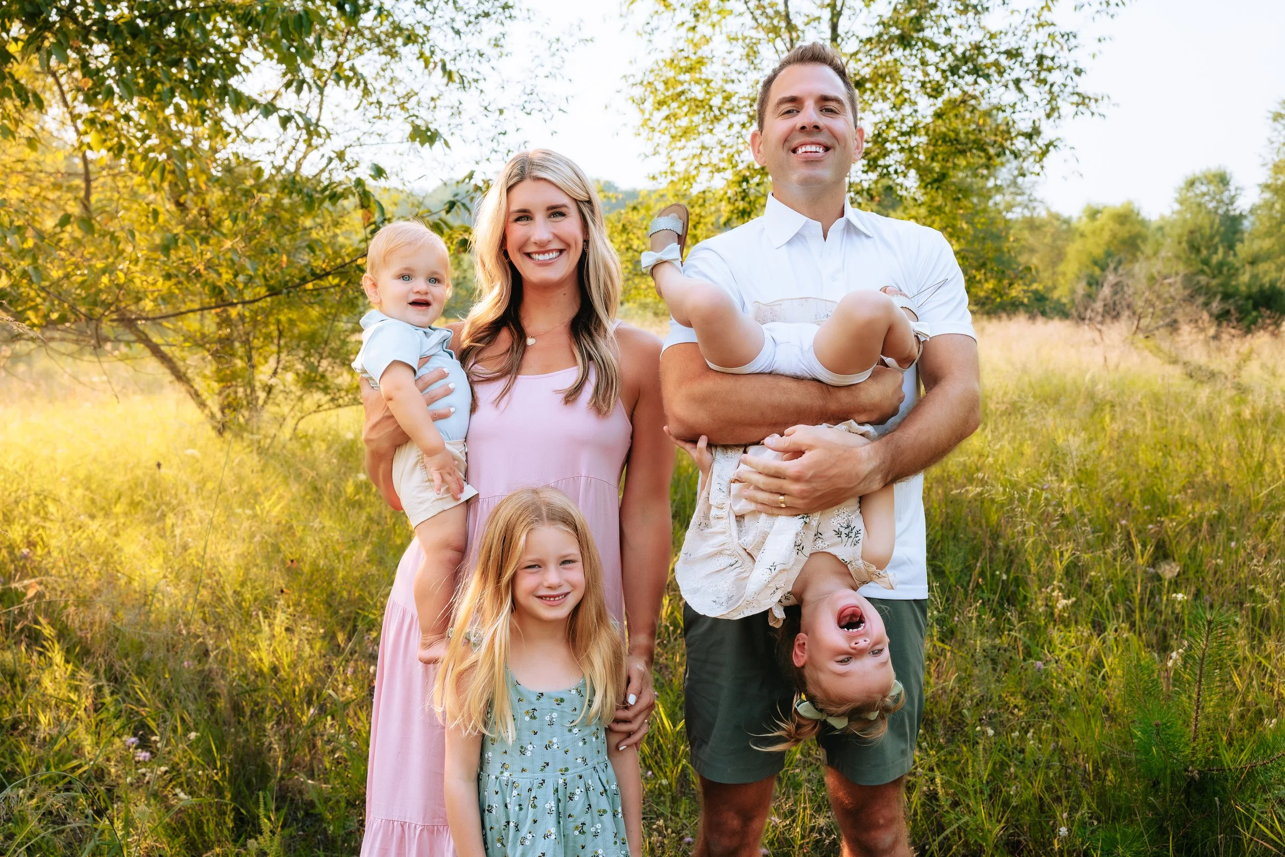A family of five outdoors in a grassy field with trees, smiling and posing for a photo. The mother and father are holding their young children, with one child hanging upside down and the other sitting on the father's shoulder.
