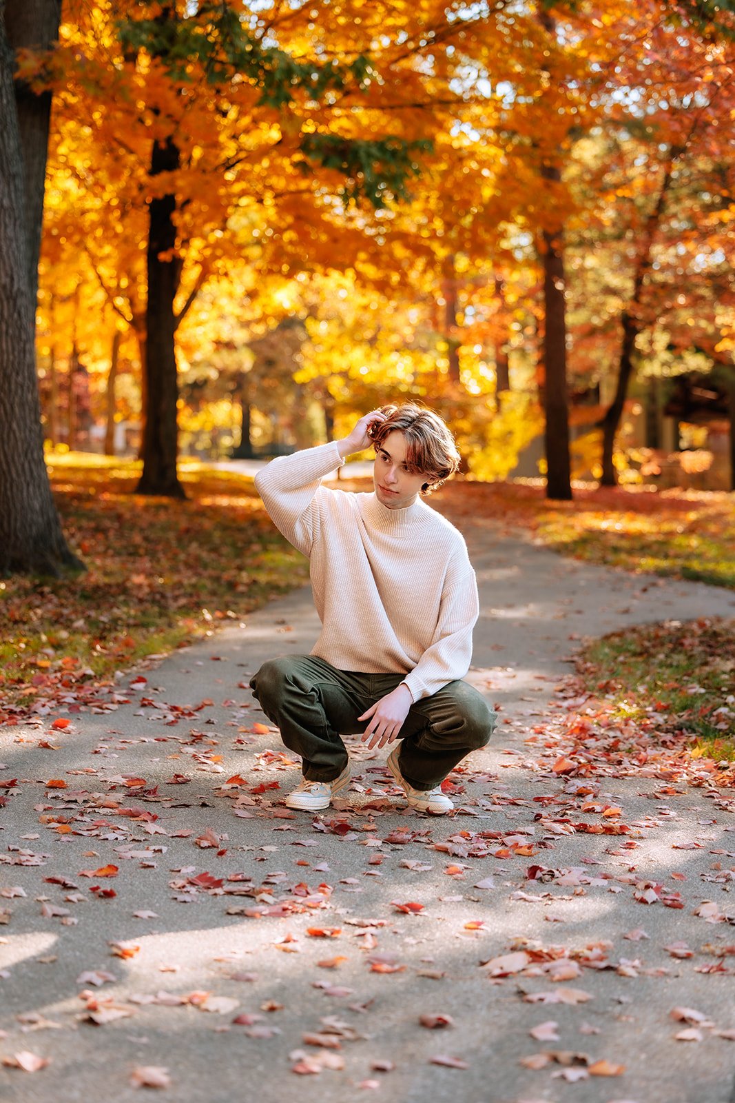 traverse city high school senior squatting on a leaf-covered path in a park with vibrant autumn foliage.