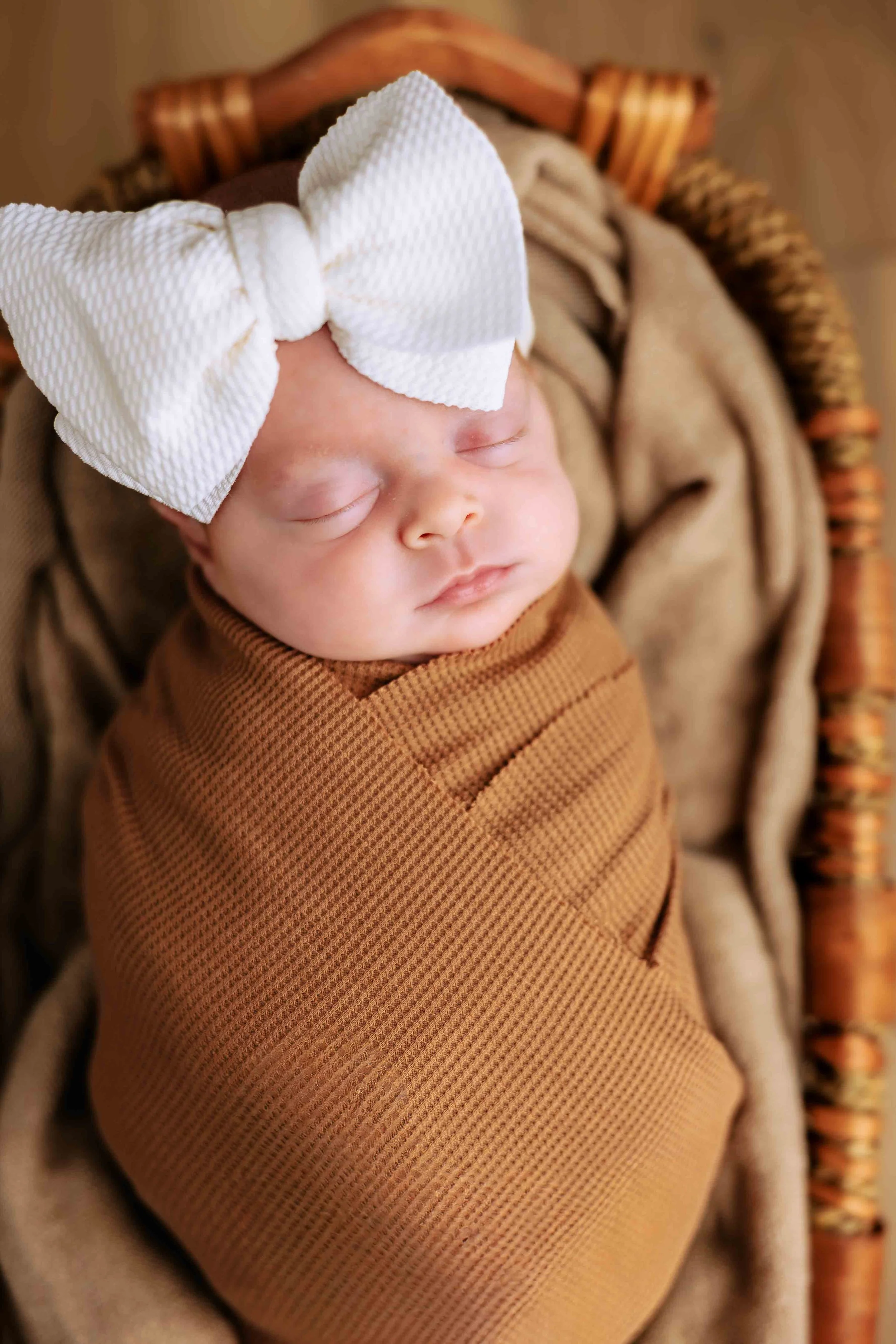 Close-up of a sleeping newborn baby wrapped in a brown blanket, wearing a large white bow on their head, lying in a wicker basket.