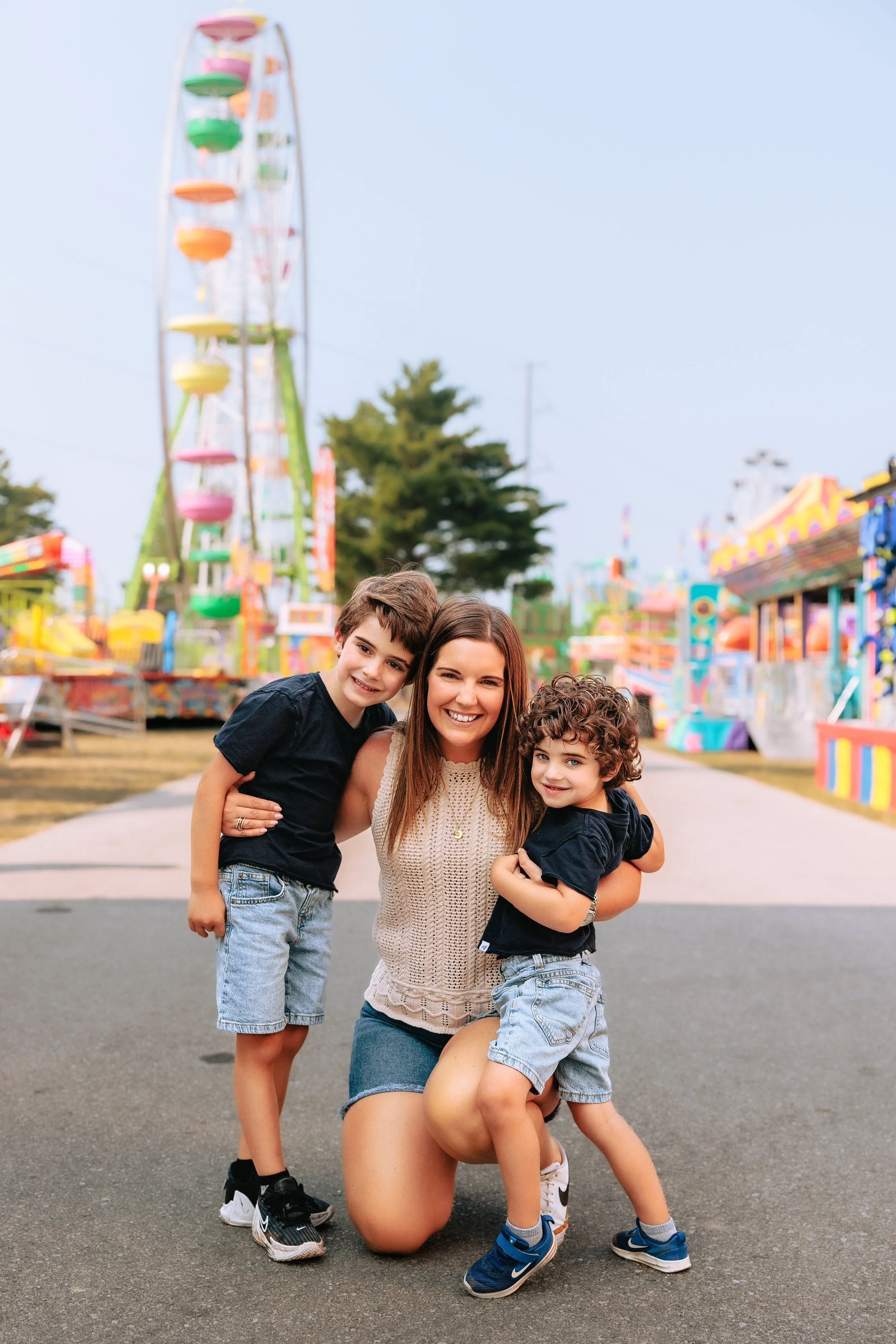 A woman with two young boys at a colorful carnival, with a Ferris wheel and amusement rides in the background.