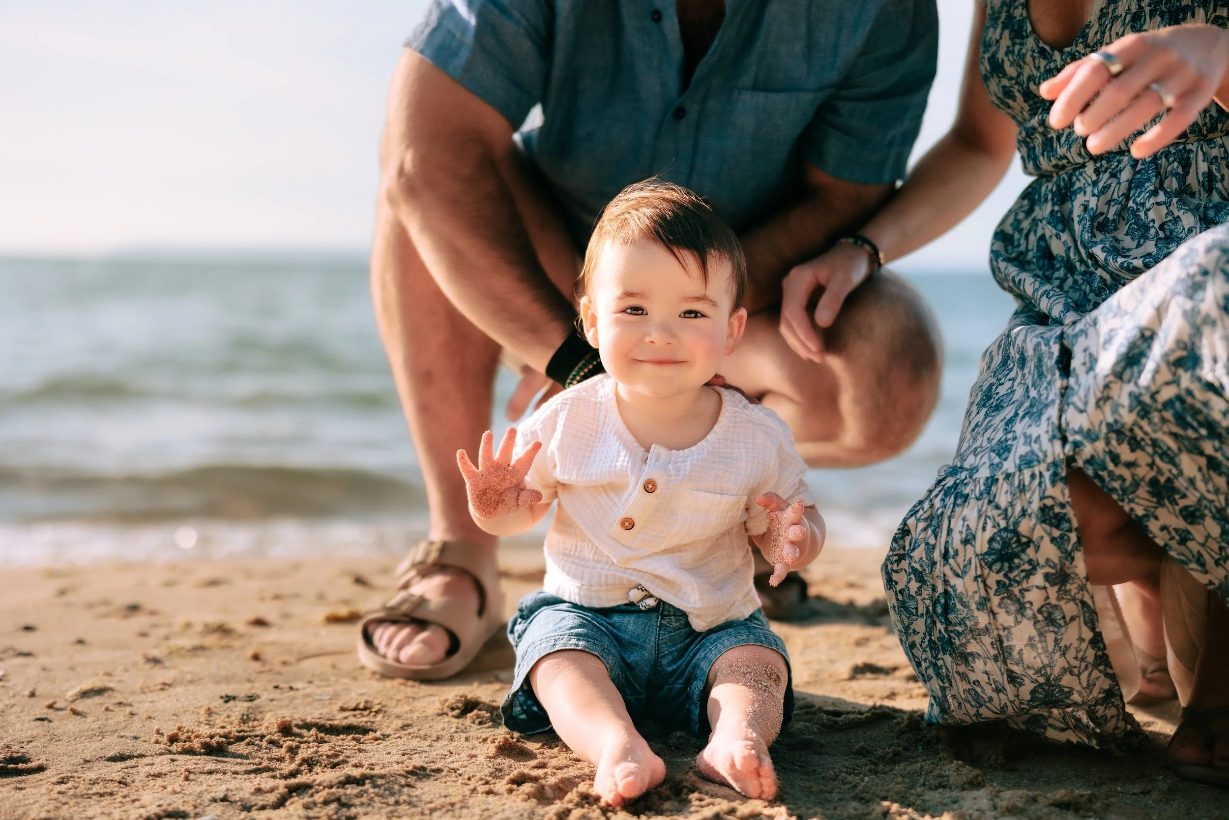 A smiling young child sitting on a sandy beach with sand on hands and legs, surrounded by three adults who are crouching nearby, with a lake and sky in the background.