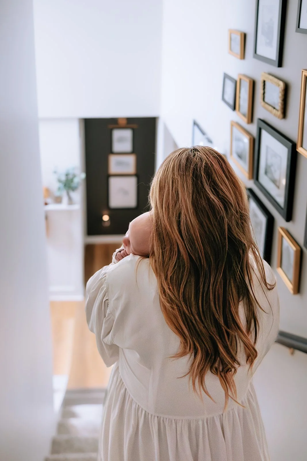 A woman with long, wavy brown hair holding a sleeping baby while standing on a staircase in a home, with a gallery wall of framed pictures on the wall beside her.