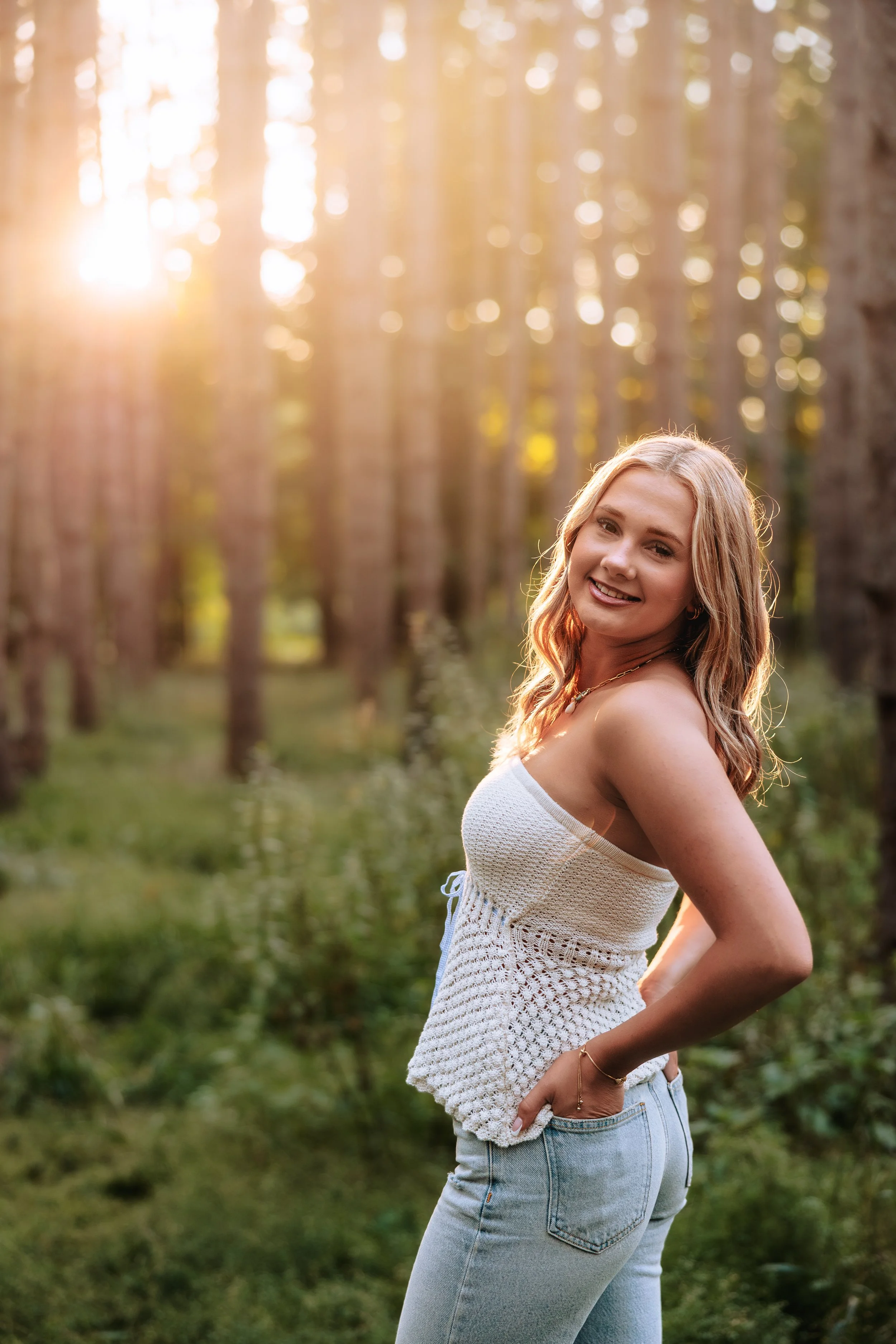 A young woman with blonde hair smiling and posing outdoors in a wooded area during sunset, wearing a white sleeveless knitted top and light blue jeans.